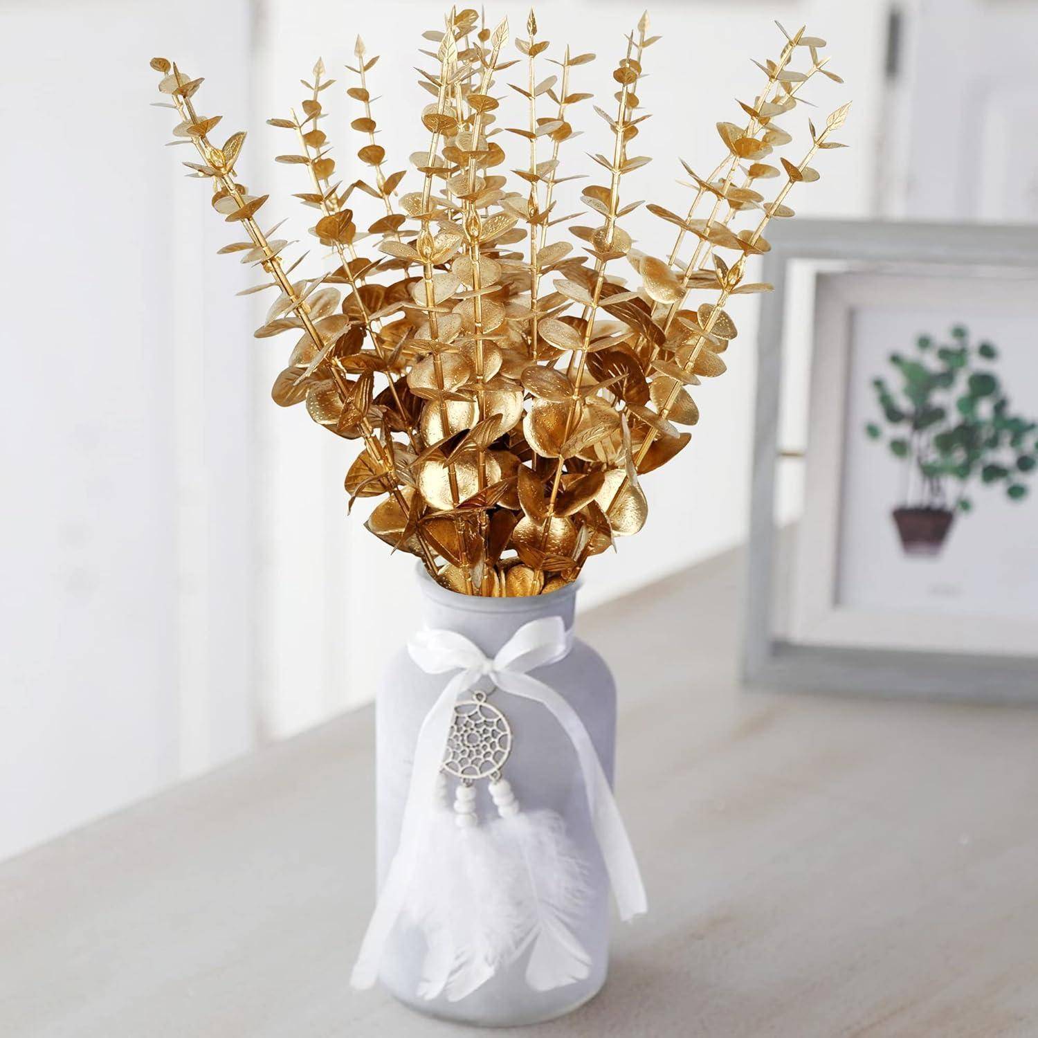 Eine weiße Vase mit goldenen, metallisch aussehenden Blättern steht auf einem Tisch. Sie ist mit einer dekorativen weißen Quaste mit Federn geschmückt.