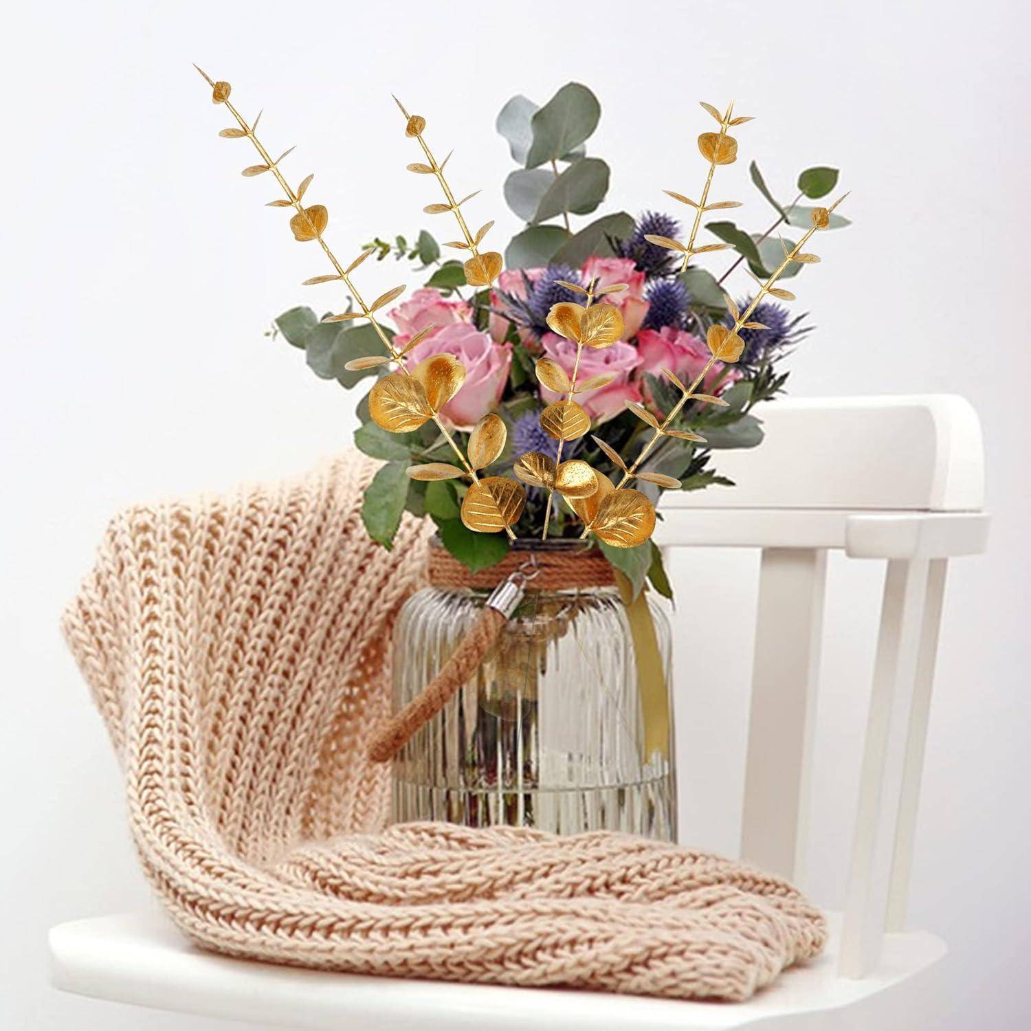 Ein Blumenstrauß in Rosa, Blau und Gold steht in einer Glasvase auf einem weißen Stuhl, begleitet von einer beigen gestrickten Decke.