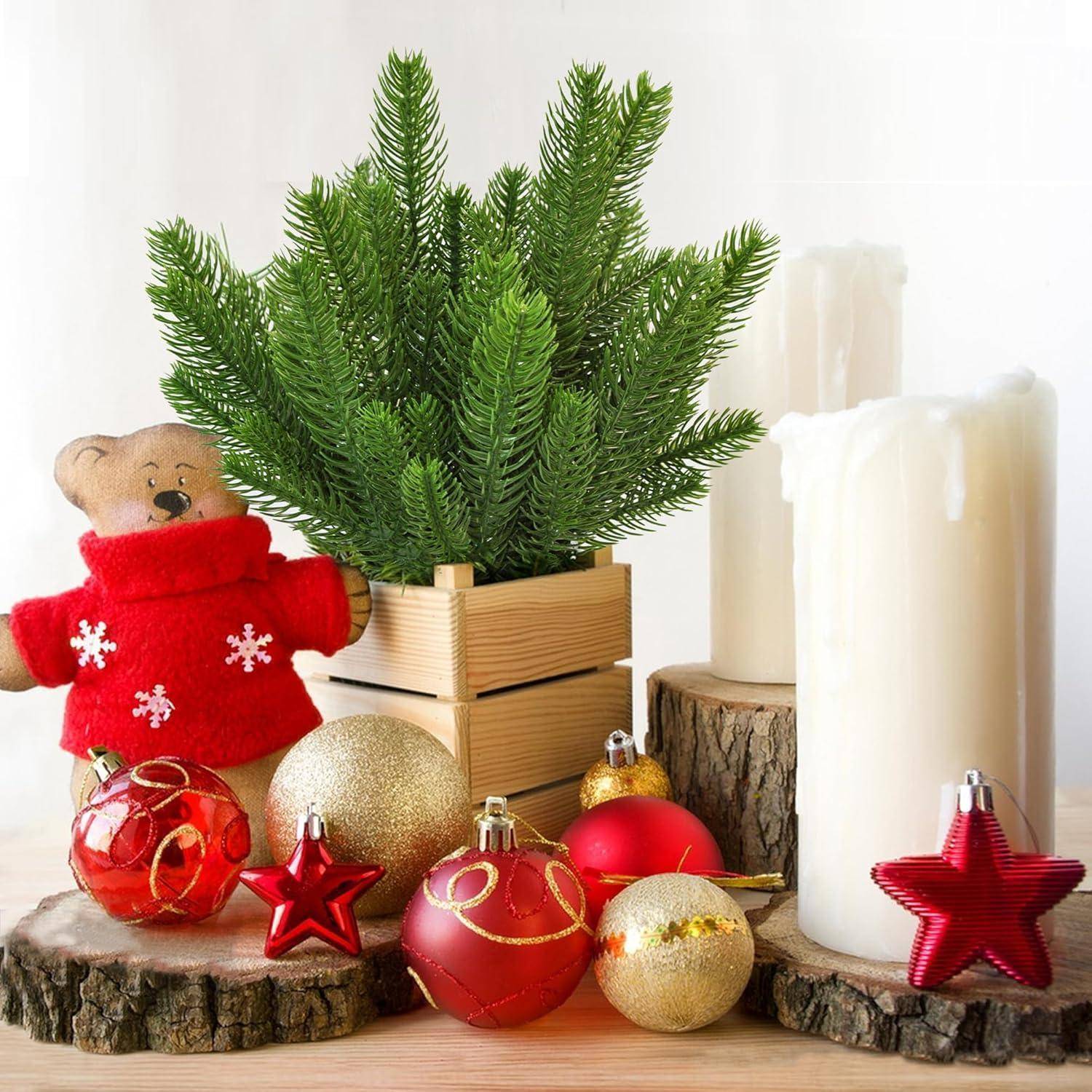 Eine festliche Anordnung mit einem kleinen Kiefernbaum in einer Holzkiste, einem Teddybären in einem roten Pullover, Kerzen sowie roten und goldenen Weihnachtsschmuck.