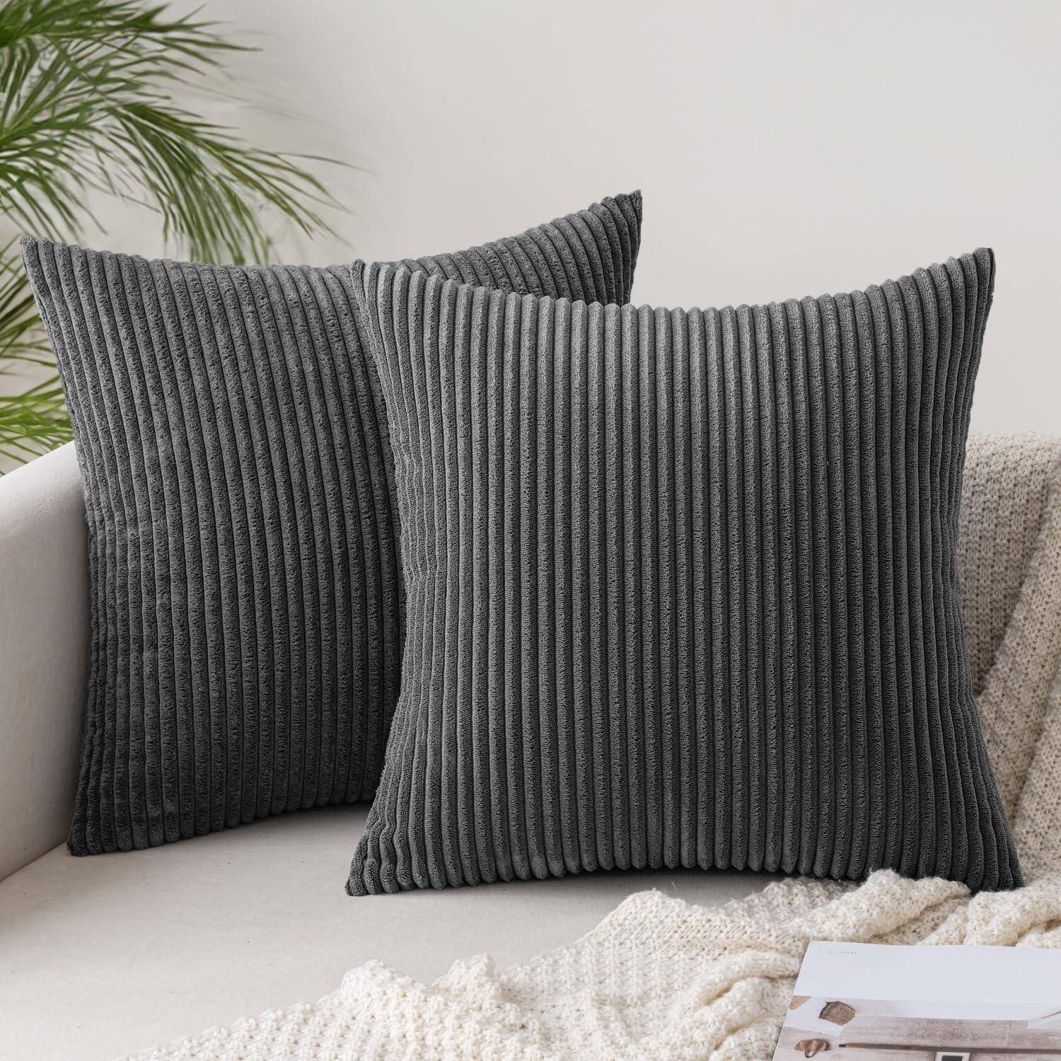 Zwei graue gerippte Zierkissen auf einem hellbeigen Sofa, mit grünen Pflanzenlättern im Hintergrund, die eine gemütliche und moderne Atmosphäre schaffen.