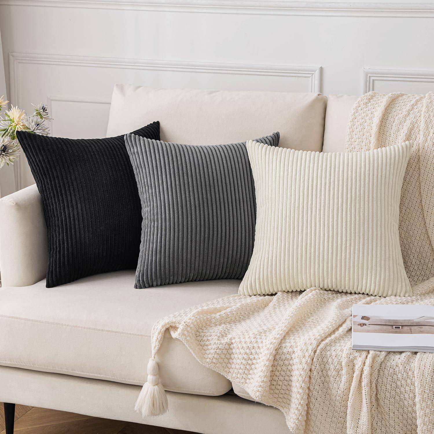 Ein beige-grauer Sofa mit drei gerippten Kissen. Die Kissen sind schwarz, grau und creme, von links nach rechts angeordnet. Eine beige Decke ist über das Sofa gebreitet.