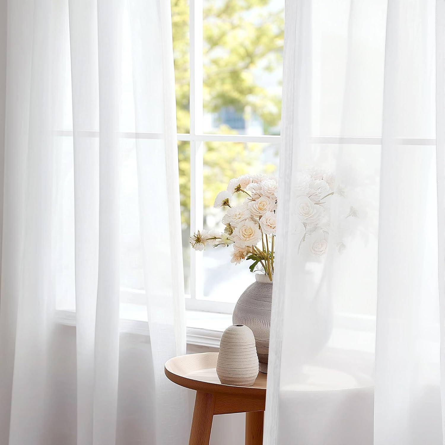 Ein kleiner Holztisch mit einer Vase voller weißer Blumen steht neben einem Fenster mit zarten weißen Gardinen, die weiches natürliches Licht in den Raum eindringen lassen.