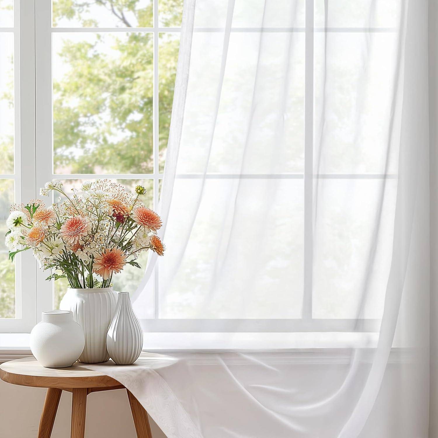 Eine Vase mit orangen und weißen Blumen auf einem kleinen Holztisch neben einem Fenster mit transparenten Gardinen, die natürliches Licht in den Raum eindringen lassen.