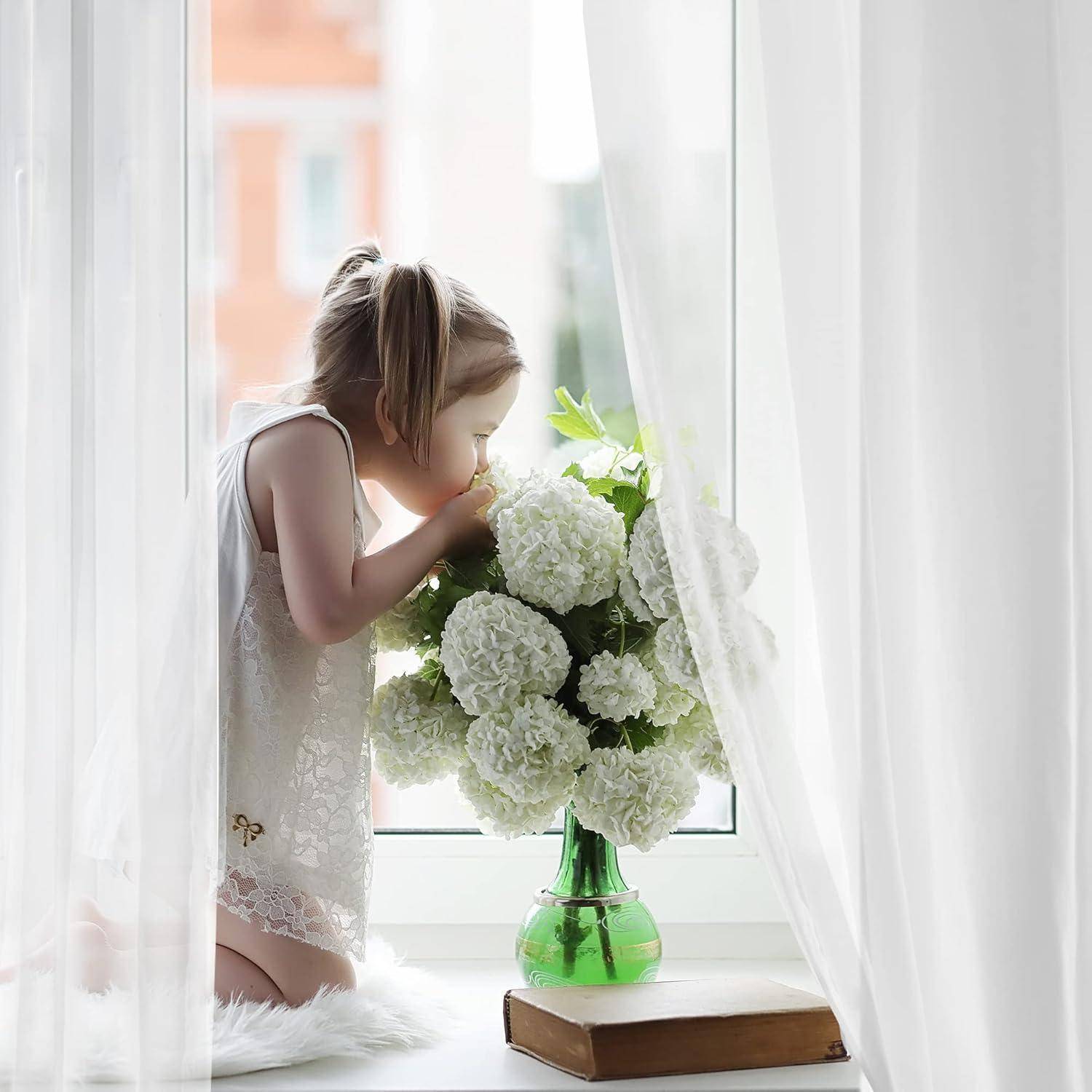 Ein junges Mädchen kniet auf der Fensterbank und riecht an einem Blumenstrauß aus weißen Blumen in einer grünen Vase, wobei durchsichtige Vorhänge die Szene umrahmen.