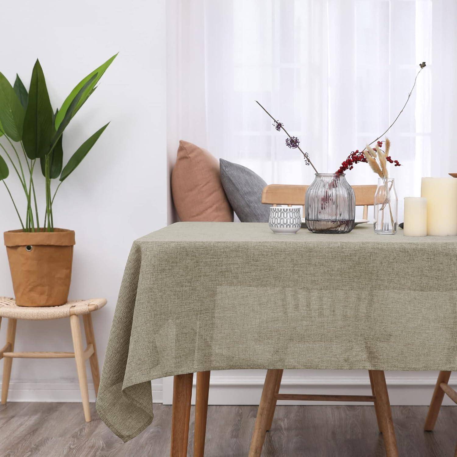 Ein Tisch mit einer neutralen Tischdecke trägt eine Vase mit getrockneten Blumen, Kerzen und ein Kissen auf einem Stuhl in einem gemütlichen, minimalistischen Raum.