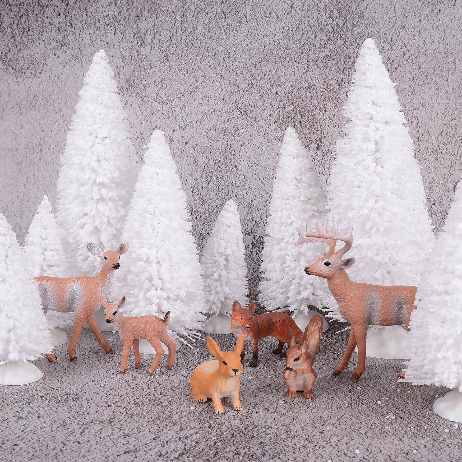 Eine Gruppe von Spieltieren, darunter Hirsche und Hasen, steht zwischen weißen, schneebedeckten Modelltannen vor einem strukturierten, grauen Hintergrund.