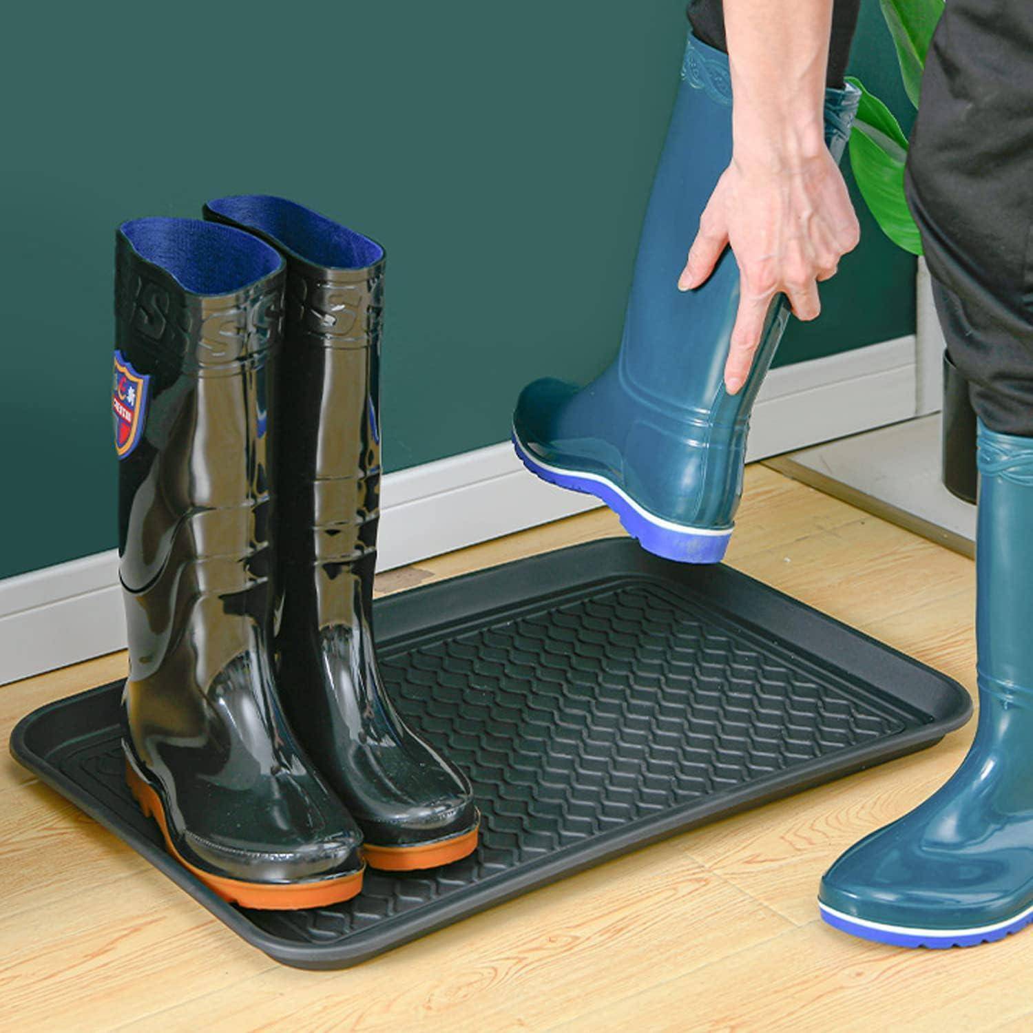 Eine Person zieht blaue glänzende Gummistiefel auf einer rechteckigen schwarzen Matte neben einem Paar schwarzer Stiefel ab, vor einer grünen Wand.