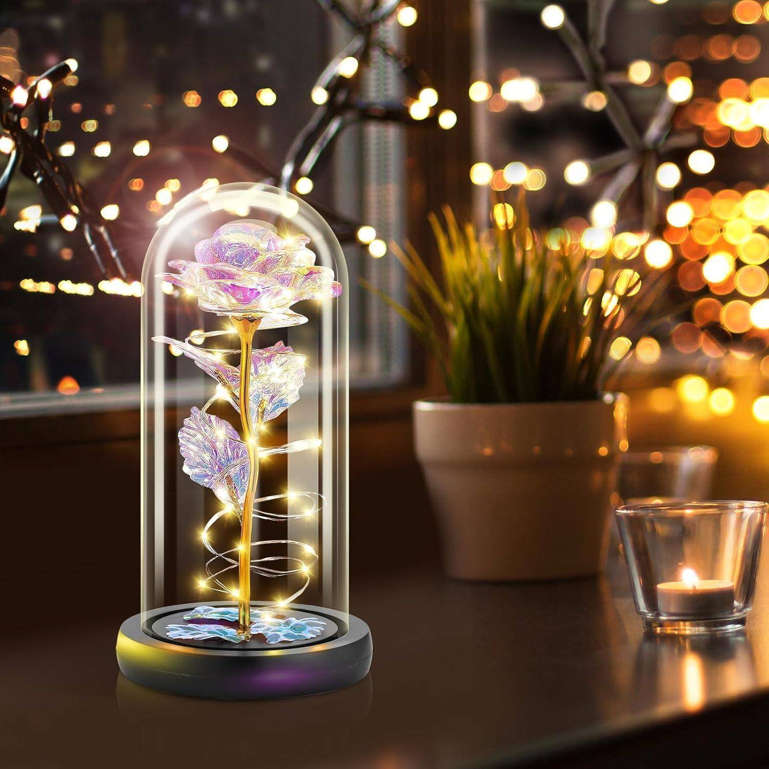 Eine leuchtende, mehrfarbige Rose, die in einer Glaskuppel eingeschlossen ist, steht auf einem Tisch neben einem Fenster, mit warmen Lichterketten und einer Pflanze im Hintergrund.