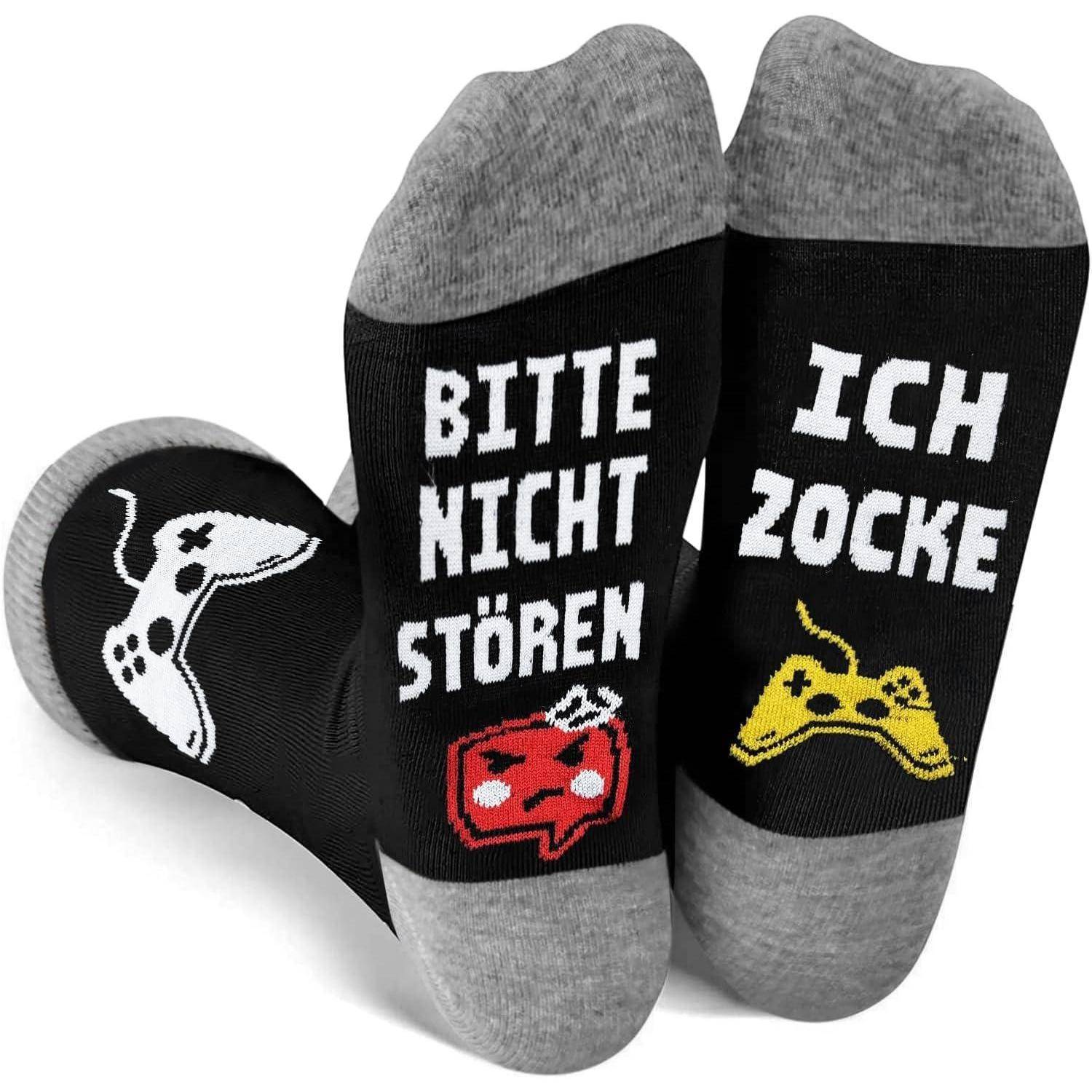 Zwei schwarze Socken mit grauen Zehenteilen und Fersen. Der linke Socken trägt die Aufschrift 'Bitte nicht stören' und zeigt einen weißen Gamecontroller. Der rechte Socken trägt die Aufschrift 'Ich zocke' mit einem gelben Controller-Symbol.