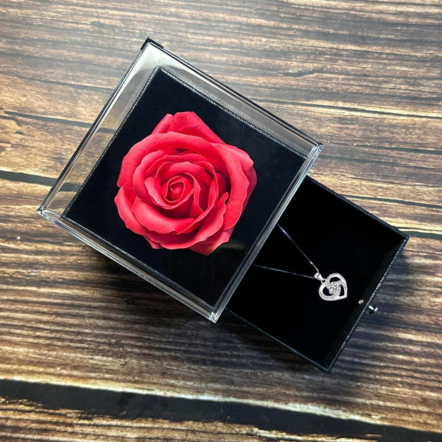 Eine rote Rose, die in einer durchsichtigen Box neben einer silbernen, herzförmigen Halskette mit einem kleinen Kristallanhänger auf einer Holzoberfläche ausgestellt ist.