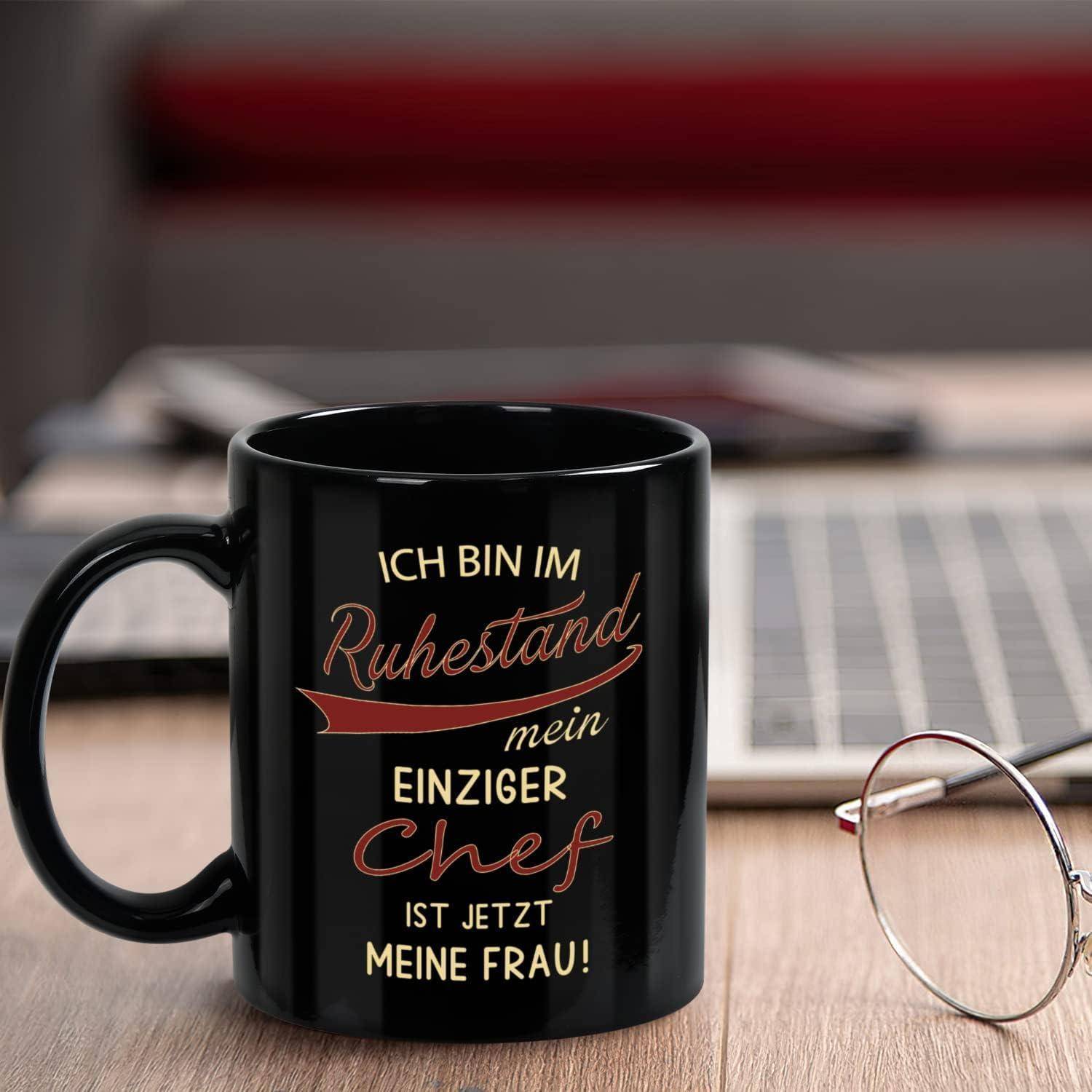 Eine schwarze Tasse mit deutschem Text: 'Ich bin im Ruhestand. Mein einziger Chef ist jetzt meine Frau!' Im Hintergrund unscharf Brille und Tastatur.