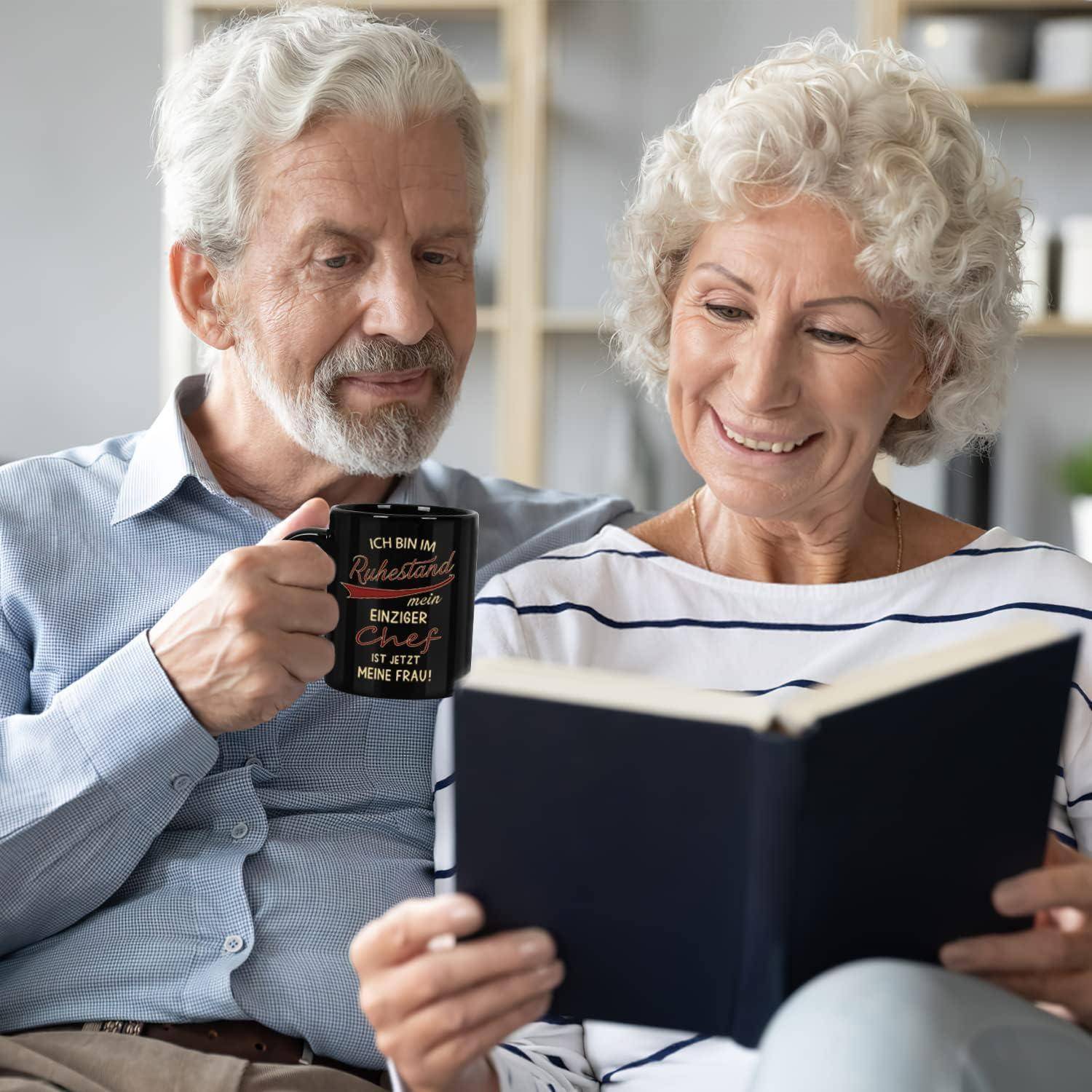 Ein älteres Ehepaar sitzt zusammen, wobei die Frau ein Buch liest und der Mann eine Kaffeetasse mit einem darauf geschriebenen Zitat hält.