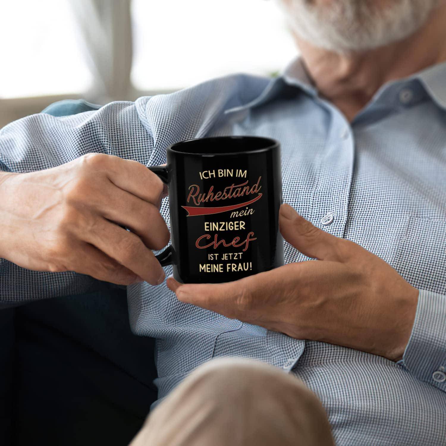 Ein älterer Mensch, der eine schwarze Tasse hält mit deutschem Text über den Ruhestand und dem Ehepartner als Chef; deutet einen humorvollen Blick auf das Leben nach der Pensionierung an.