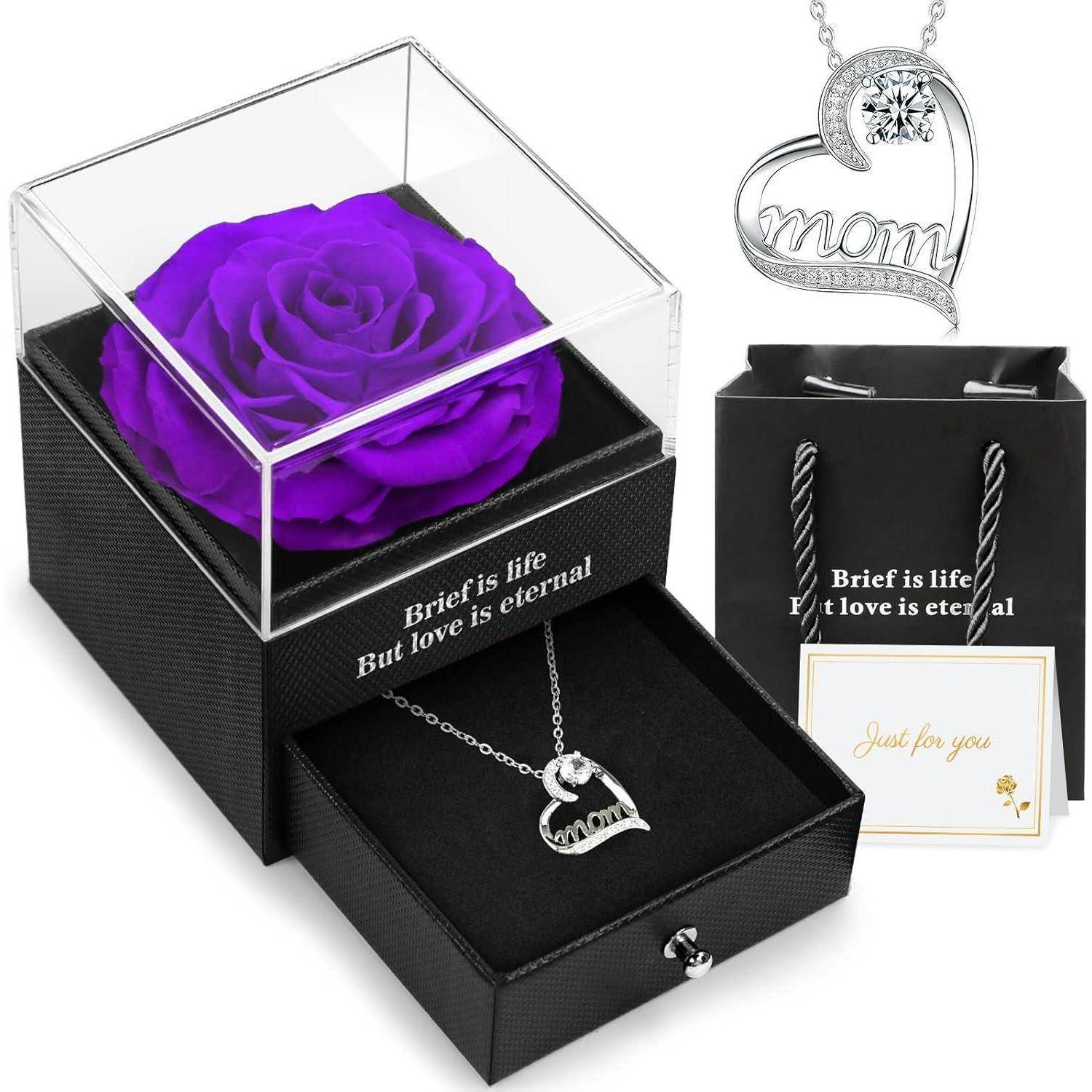 Eine lila Rose in einer durchsichtigen Box mit einer Herzkette darunter, mit der Aufschrift ‚Mama' und ‚Das Leben ist kurz, Aber die Liebe ist ewig,' neben einem Geschenkbeutel und einer Karte.