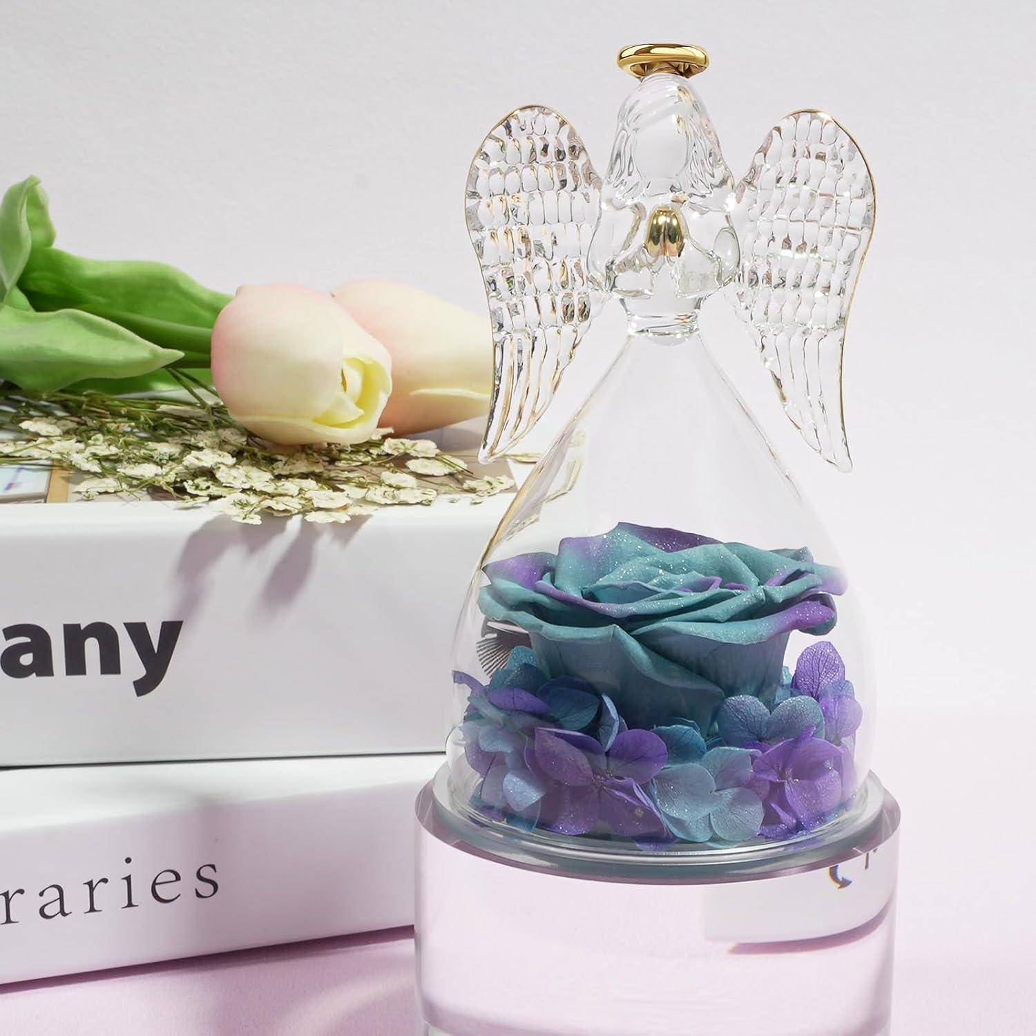 Eine Glasengelfigur mit blauen und lila konservierten Rosen im Inneren steht auf Büchern. Daneben ruhen zwei rosa Tulpen auf einer Seite.