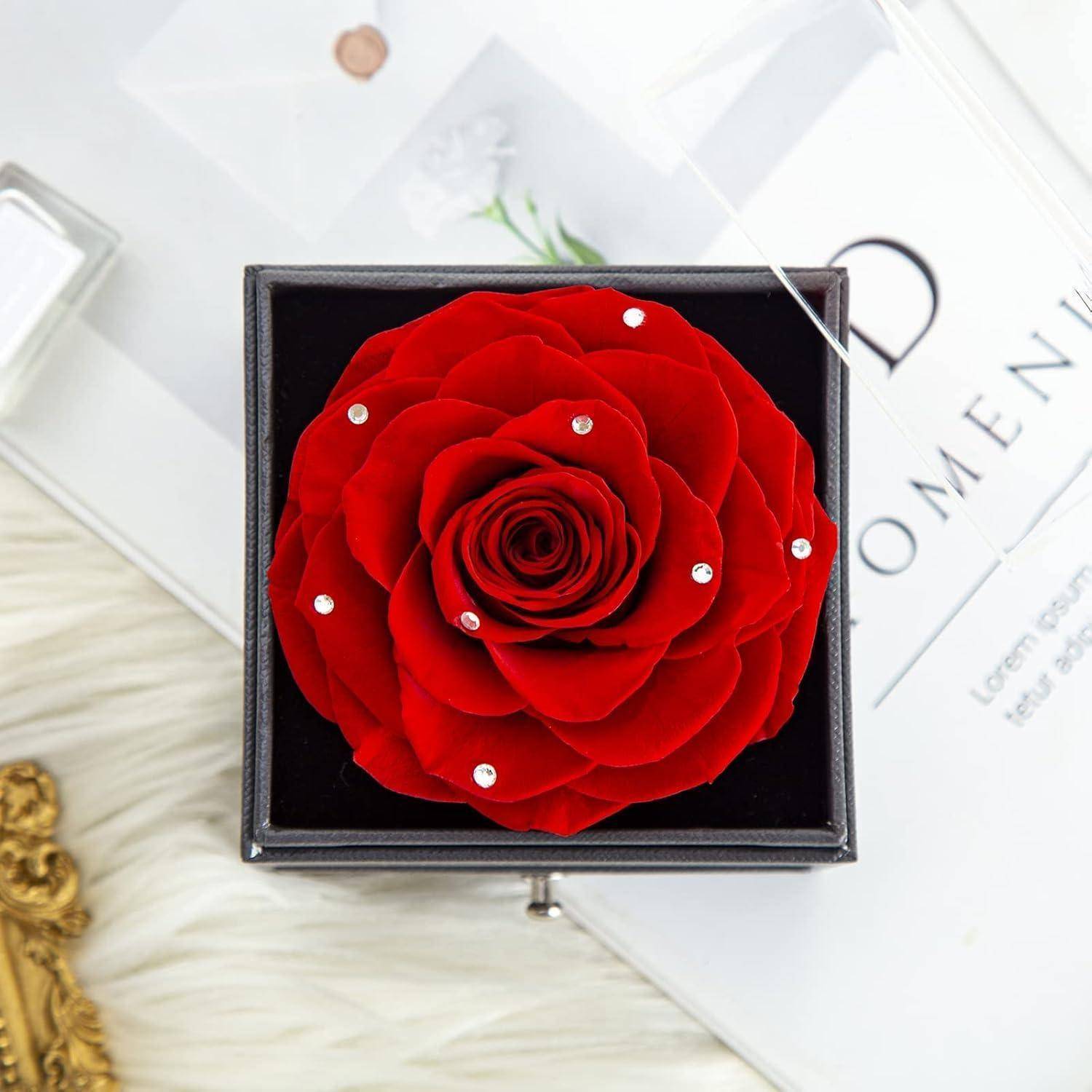 Eine lebendige rote Rose, geschmückt mit kleinen weißen Edelsteinen, wird in einer schwarzen Box präsentiert, die auf einem weißen Fell-Hintergrund mit dekorativen Elementen platziert ist.
