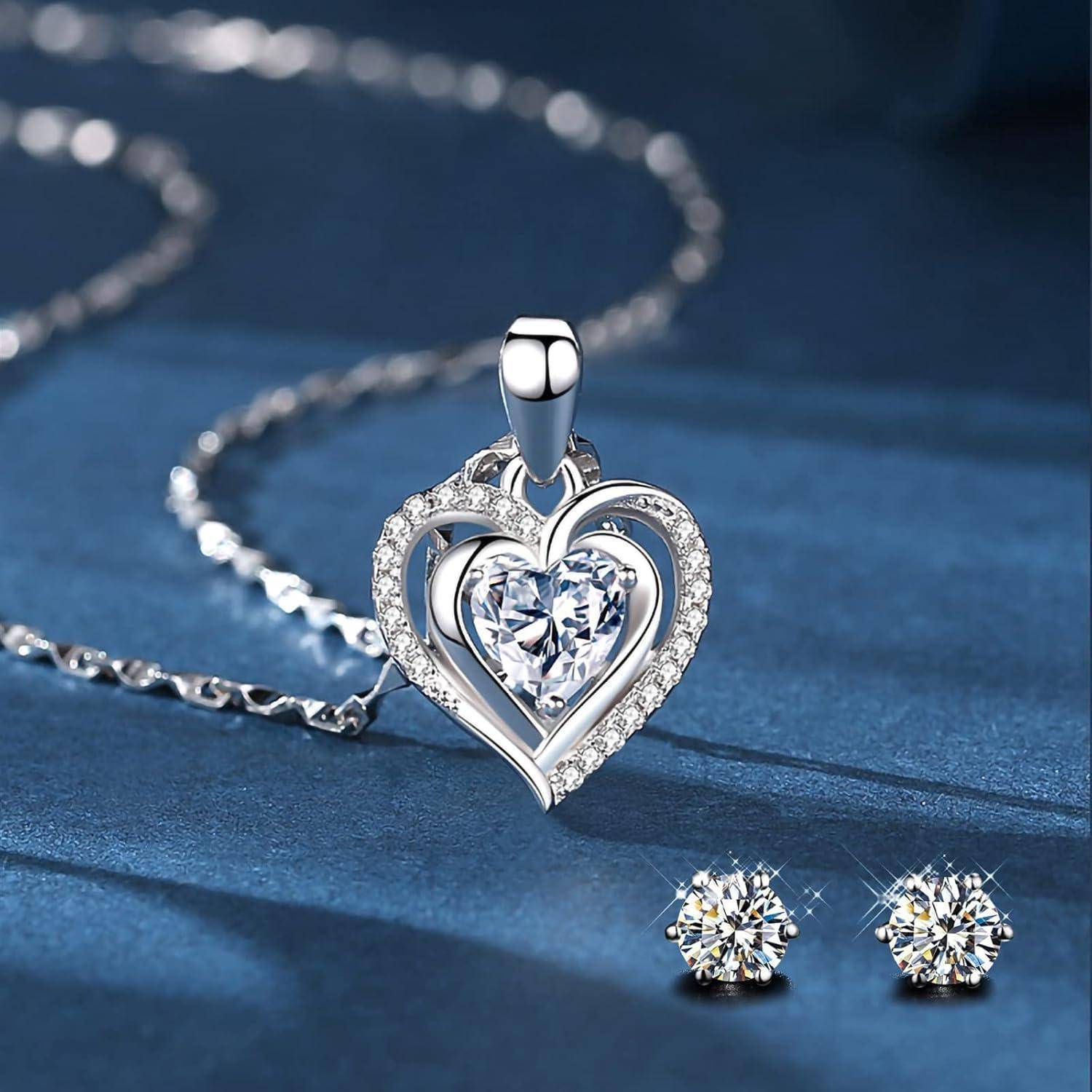 Eine silberne Halskette mit einem herzförmigen Anhänger, der einen zentralen Diamanten trägt, sowie passende Diamant-Ohrstecker vor einem blauen Hintergrund.