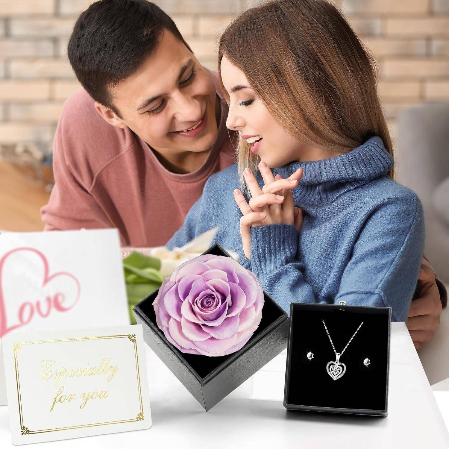 Ein Paar, das sich anlächelt, mit einer Geschenkbox im Vordergrund, die eine rosa Rose und eine herzförmige Halskette enthält.