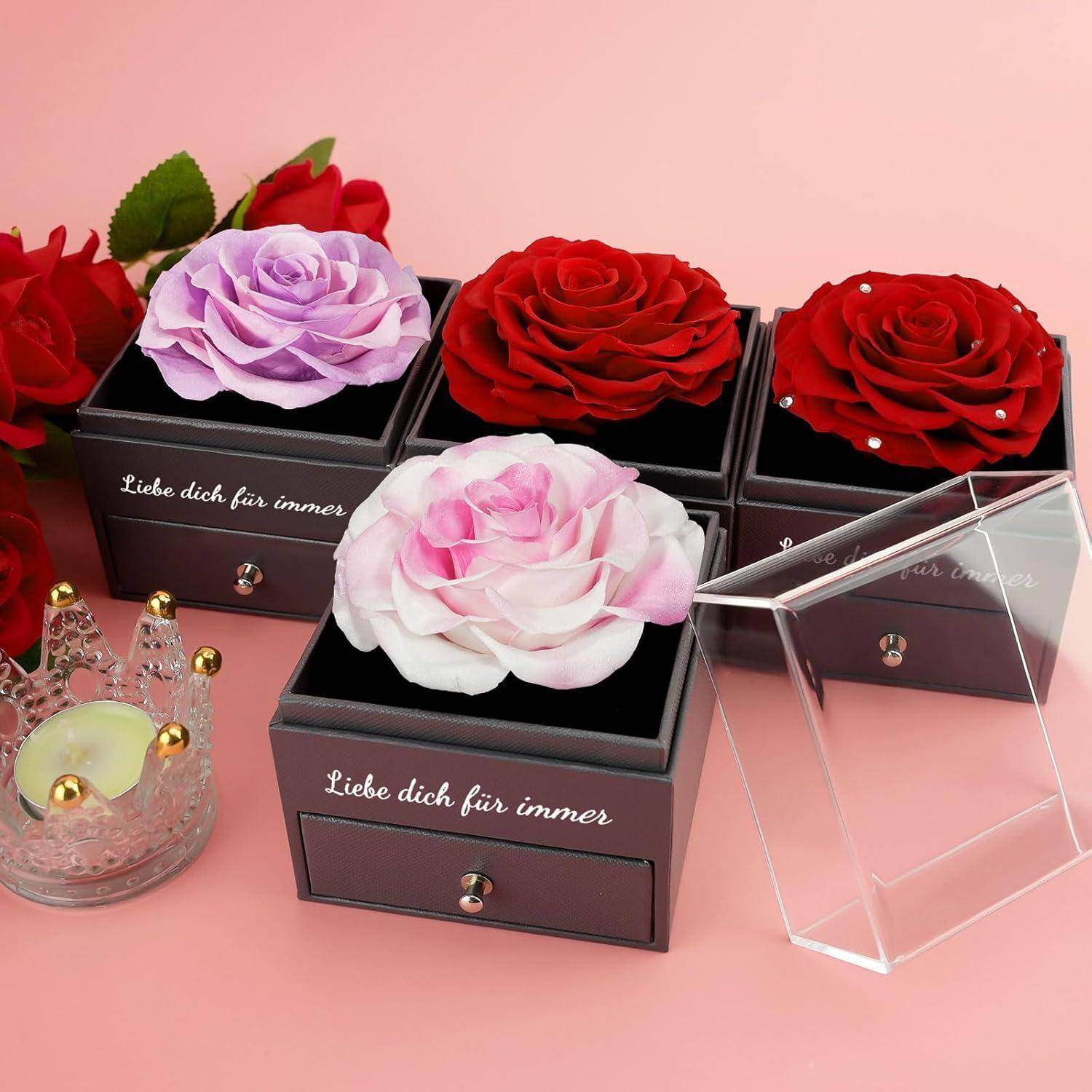 Vier dekorative Boxen, von denen jede eine konservierte Rose und den Text ‚Liebe dich für immer' enthält, vor einem rosa Hintergrund mit zusätzlichen Rosen.