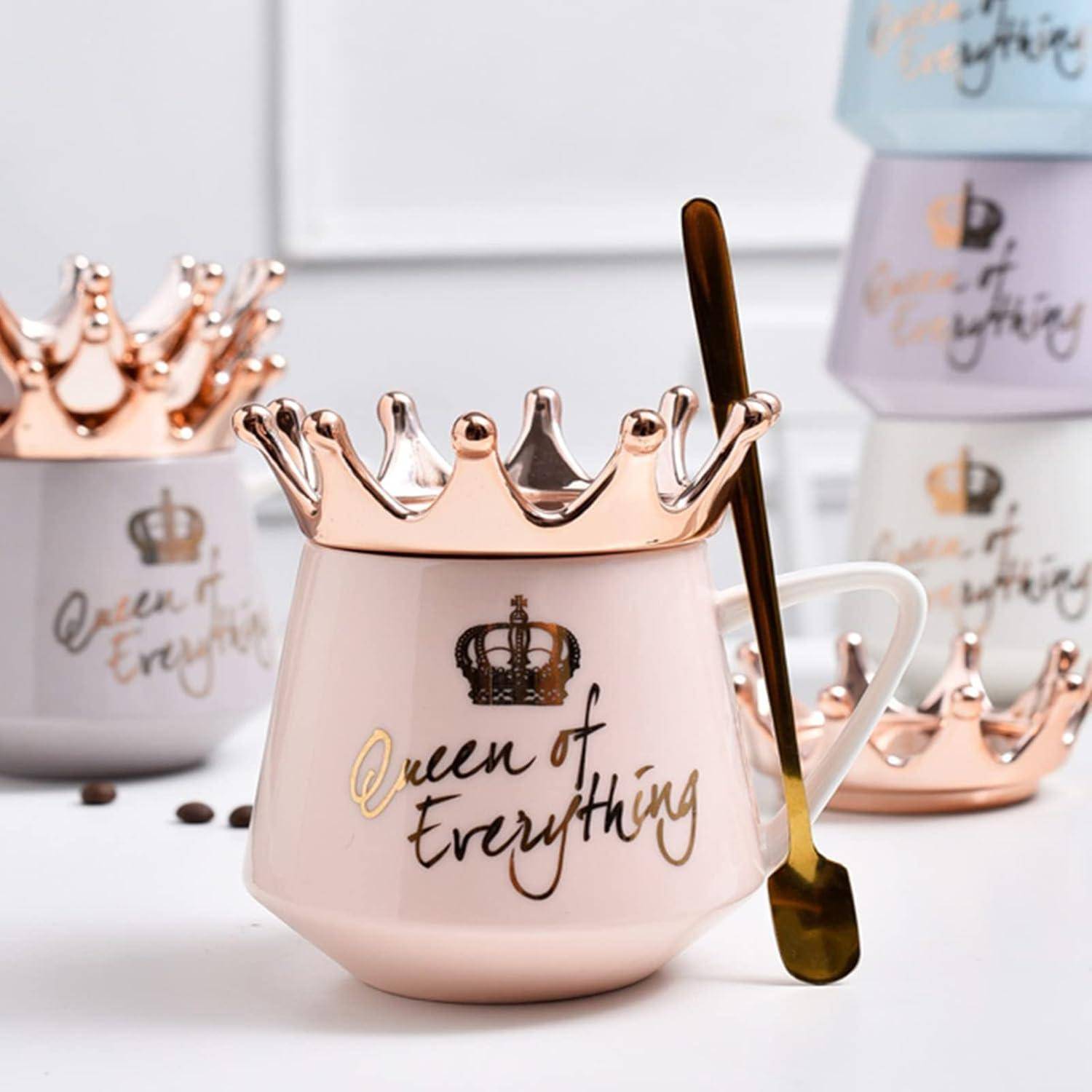 Rosa Tasse mit einer Krone als Deckel und einem goldenen Löffel daneben, auf der der Text 'Königin von Allem' zu sehen ist. Im Hintergrund zwei unscharf dargestellte Tassen.