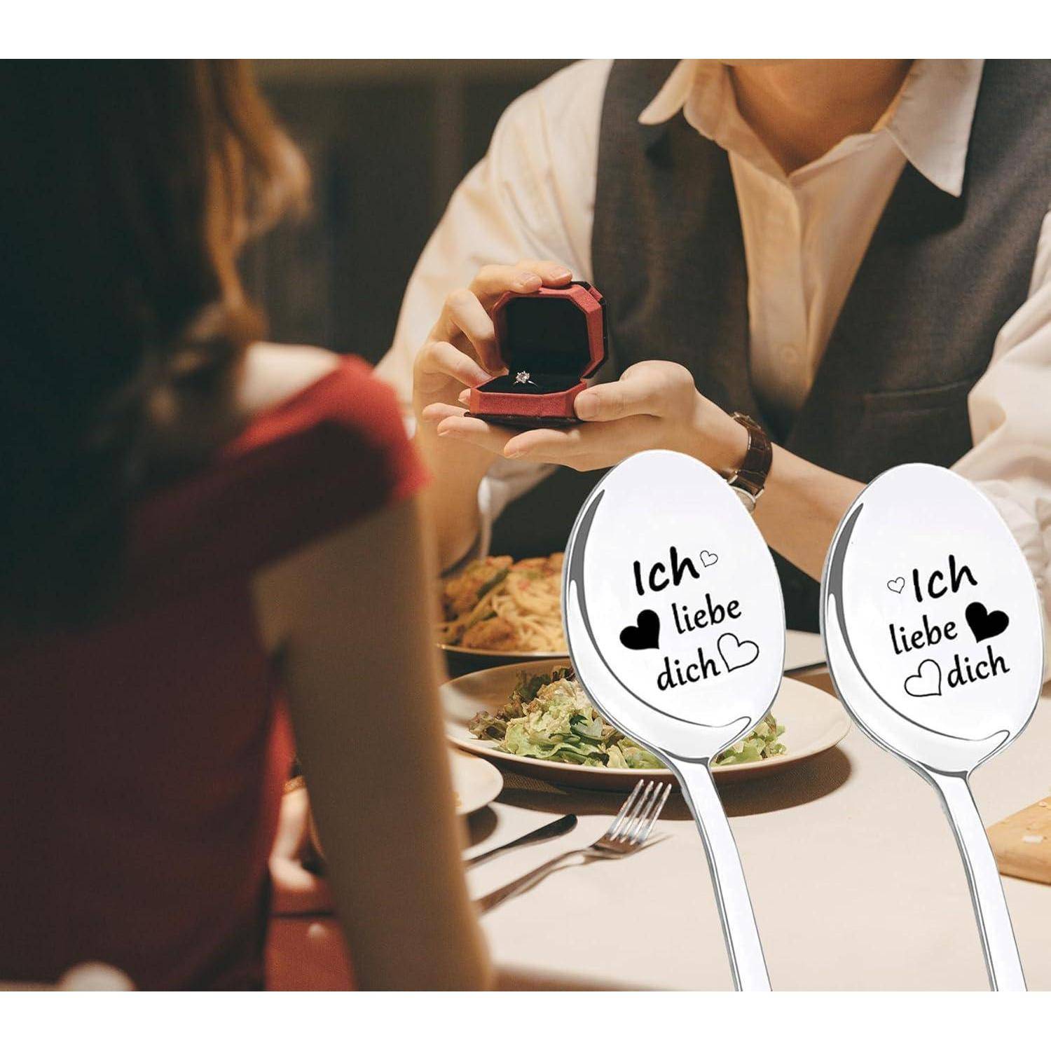 Eine Person, die an einem Esstisch einen Heiratsantrag macht und eine geöffnete Ringschachtel hält. Die Löffel im Vordergrund tragen die Aufschrift „Ich liebe dich