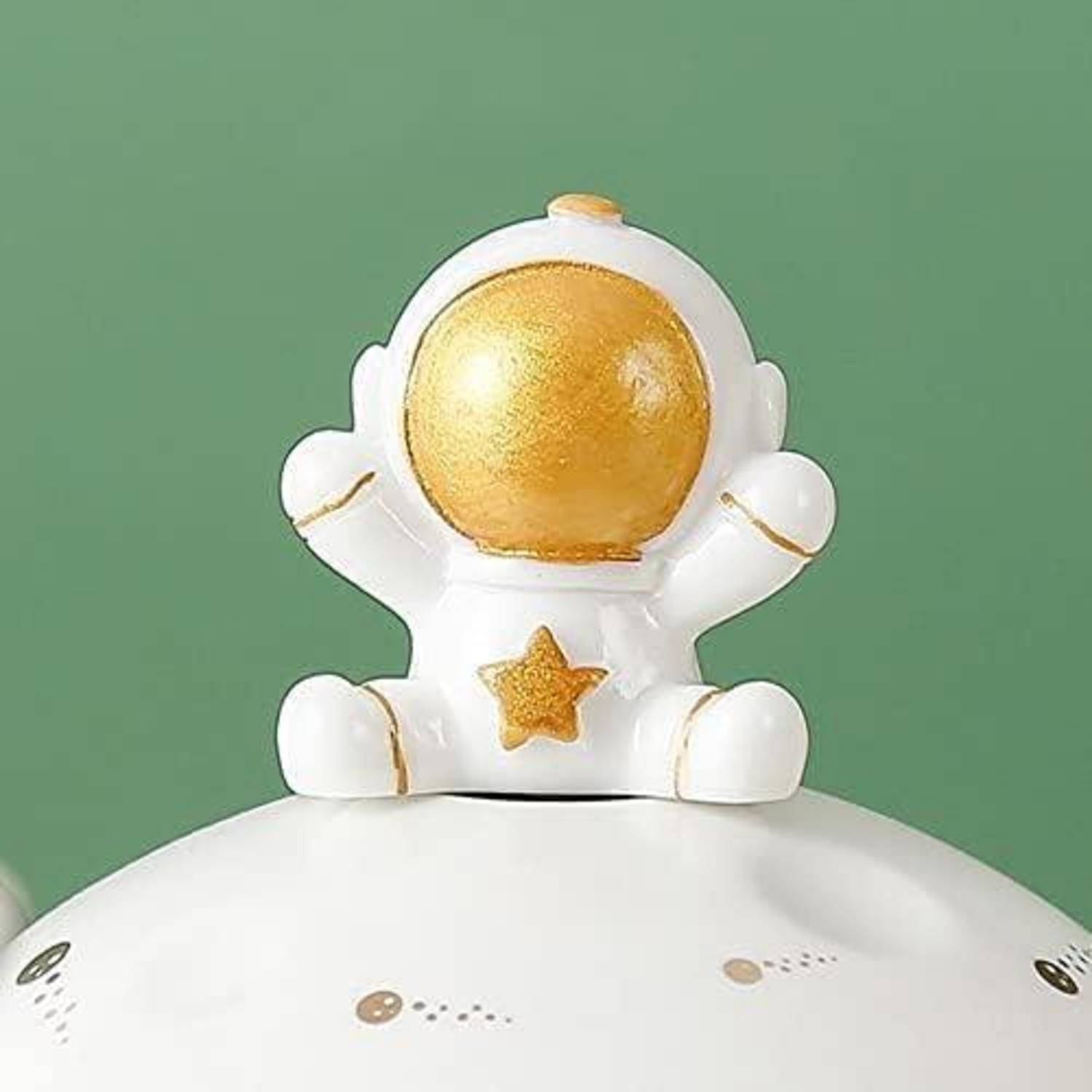 Eine kleine keramische Astronautenfigur mit goldenem Helm und Stern auf der Brust sitzt auf einer runden weißen Oberfläche vor einem grünen Hintergrund.