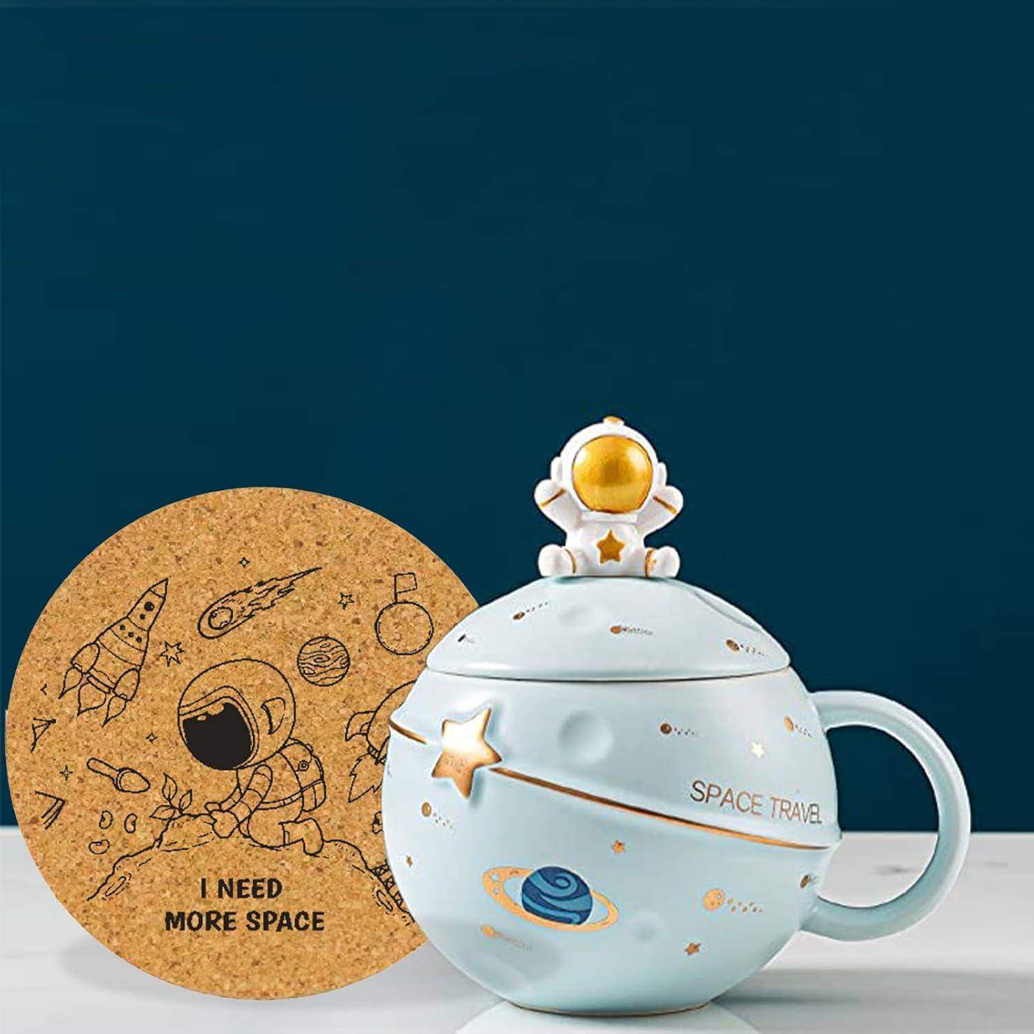 Eine dekorative blaue Tasse mit einem Stern- und Planetenmotiv, gekrönt von einer kleinen Astronautenfigur. Daneben ein Untersetzer aus Kork mit einem Astronauten und dem Spruch „Ich brauche mehr Platz