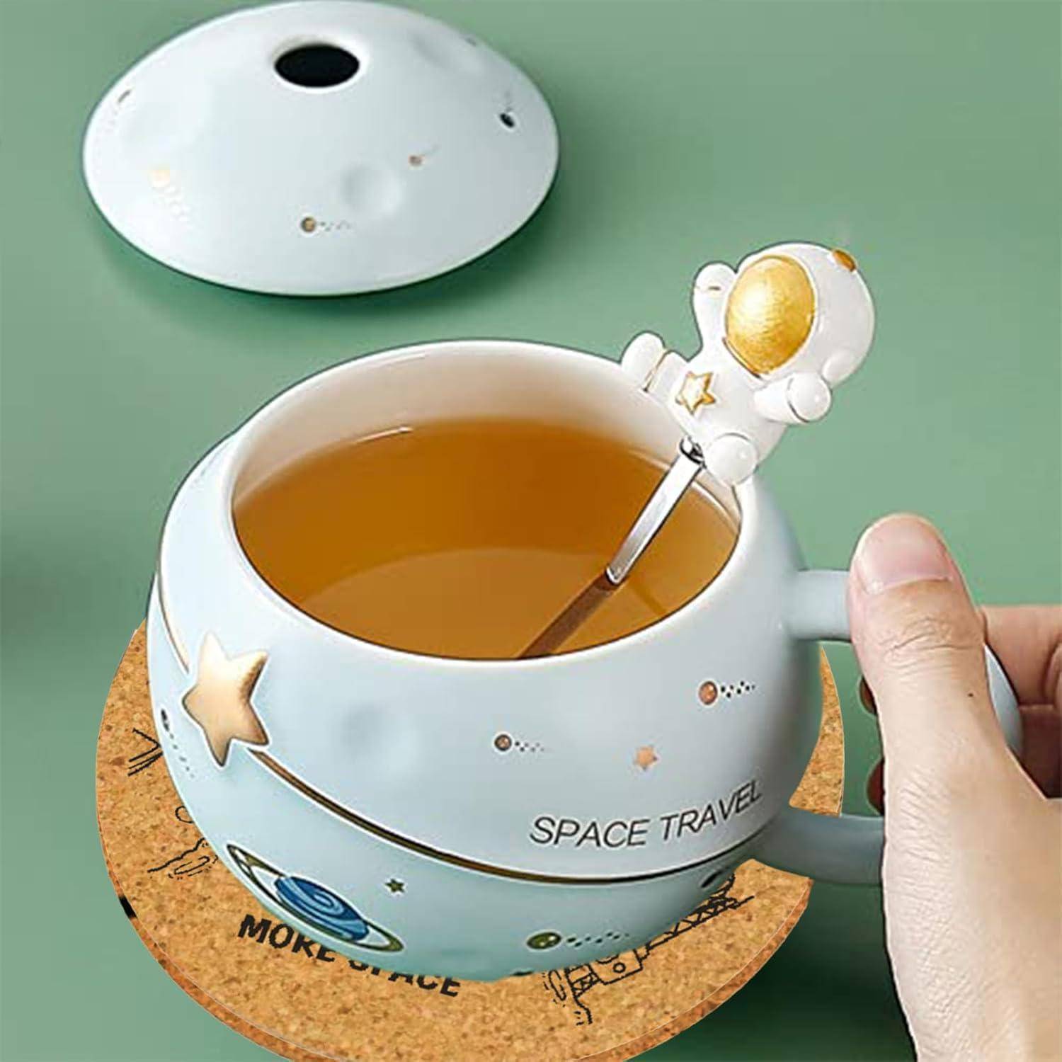 Eine Tasse mit 'Weltraumreise'-Design, Planeten und Sternen. Ein Löffel mit einem Astronautengriff liegt innen. Deckel und runder Untersetzer in der Nähe.