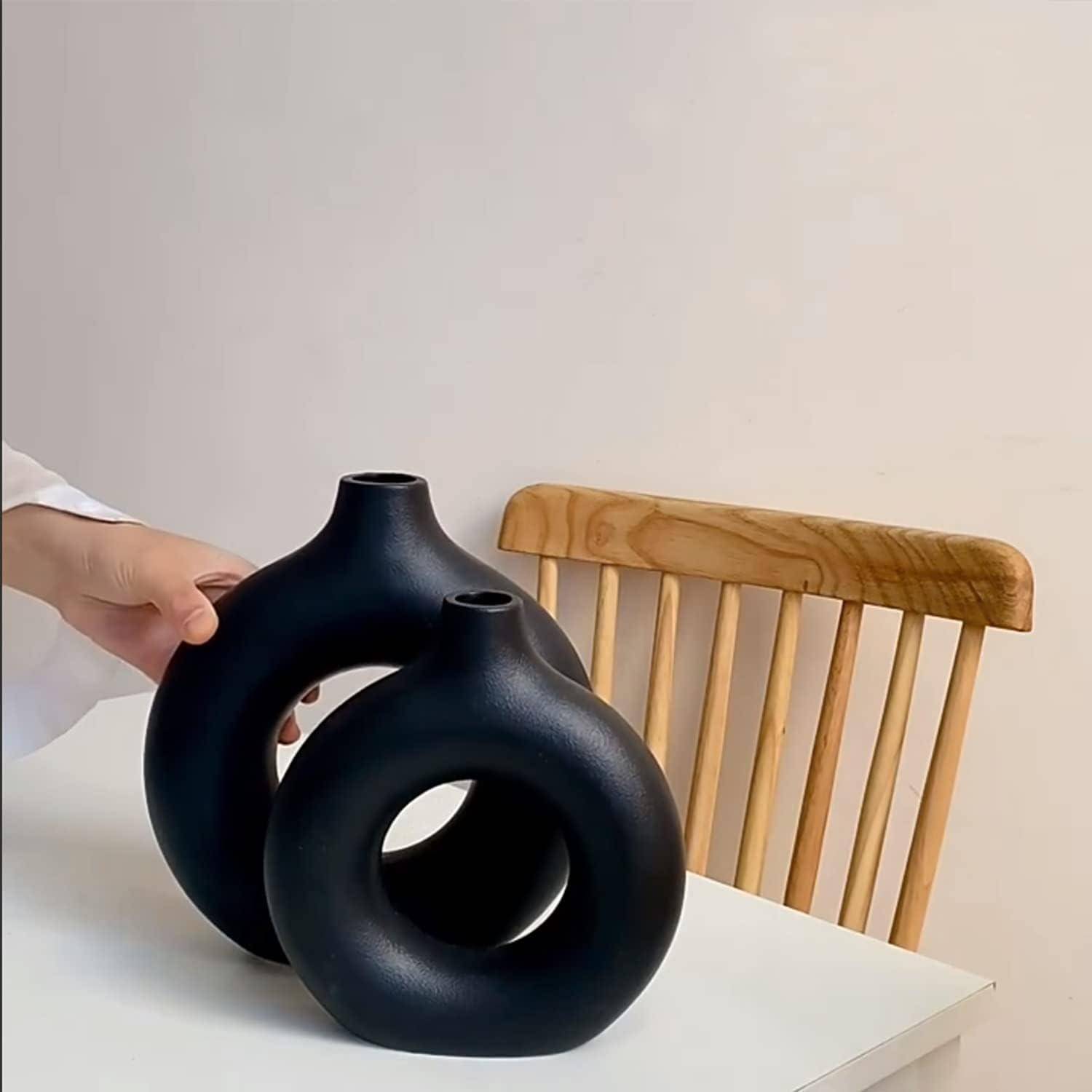 Eine Person arrangiert zwei moderne schwarze Vasen mit kreisrunden Ausschnitten auf einem weißen Tisch neben einem Holzstuhl und schafft damit eine minimalistische Einrichtungssituation.