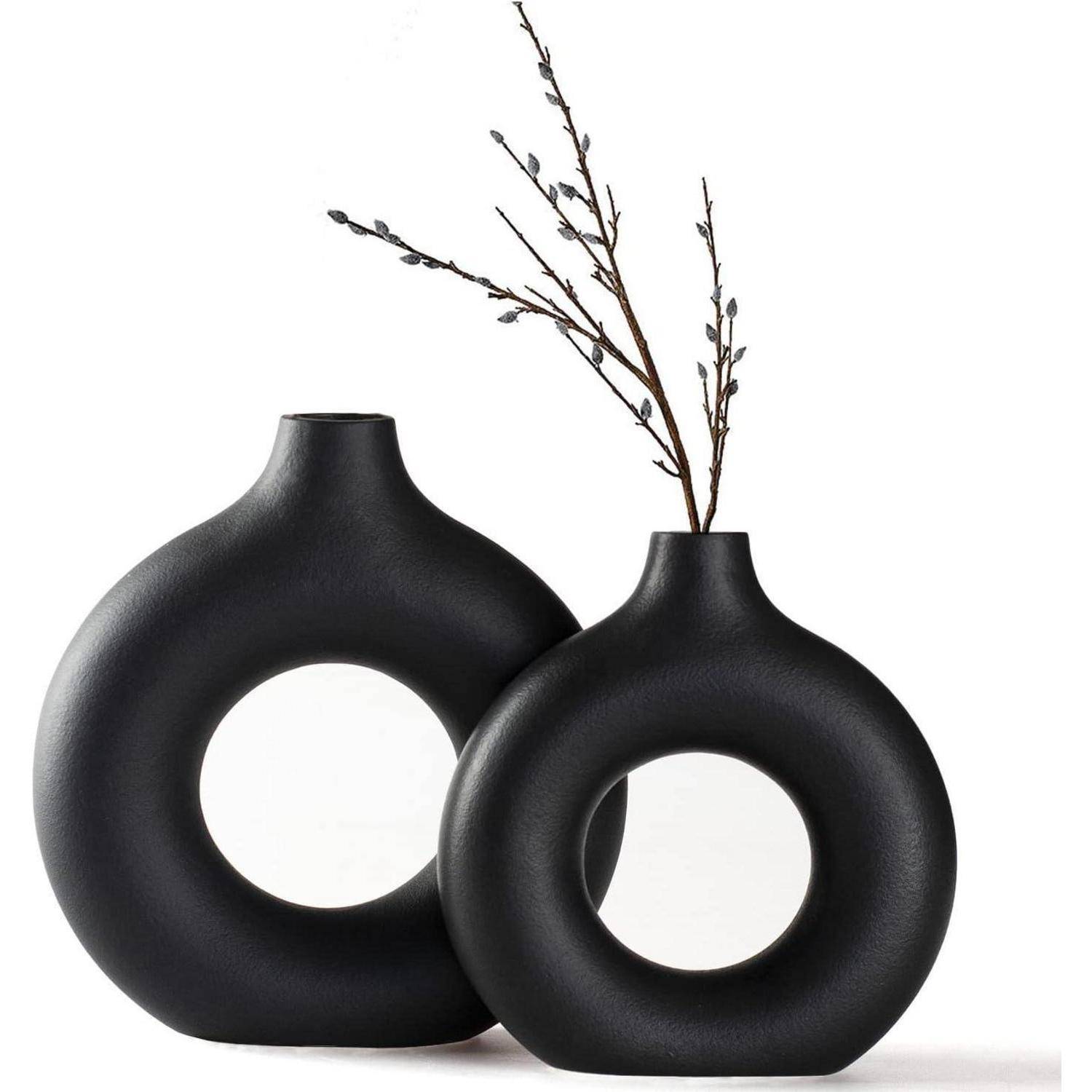 Zwei schwarze Vasen mit kreisförmigen Ausschnitten; eine kleinere Vase hält schlanke Zweige. Minimalistisches Design auf weißem Hintergrund.
