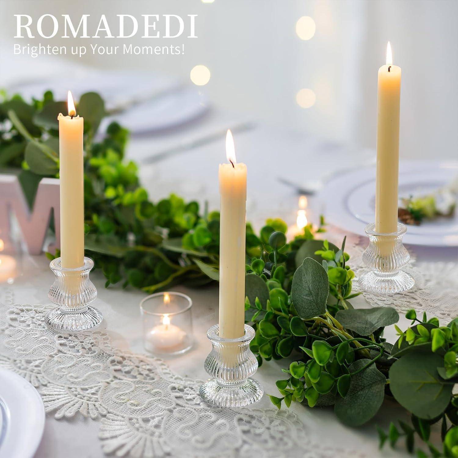 Hohe weiße Kerzen in Glashaltern auf einem Tisch mit Grünpflanzen, gedeckt für ein Abendessen. Text in der Ecke liest: 'ROMADEDI: Erhelle deine Momente!