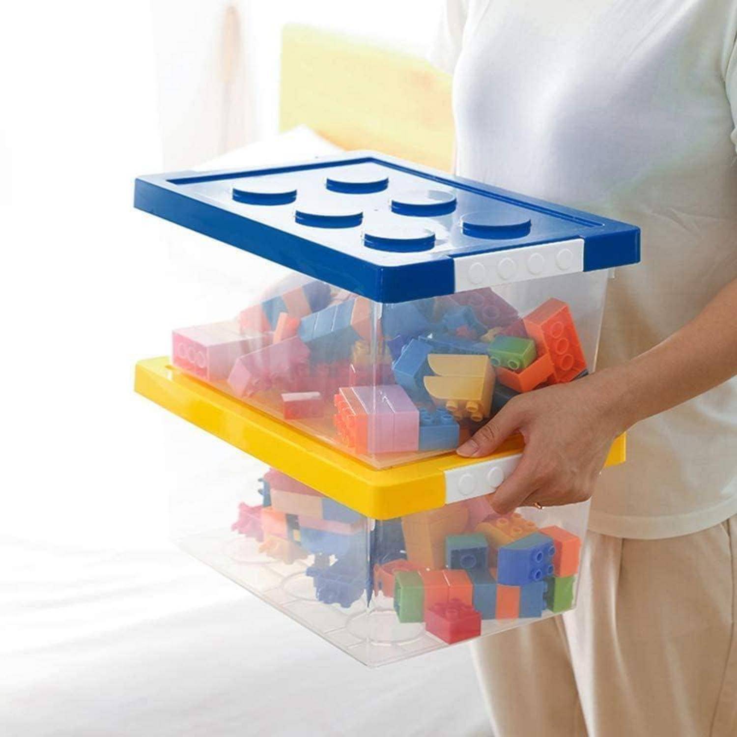 Eine Person hält einen durchsichtigen Plastikbehälter, gefüllt mit bunten Spielzeug-Bausteinen, der mit einem Deckel in Form blauer Bausteine bedeckt ist.