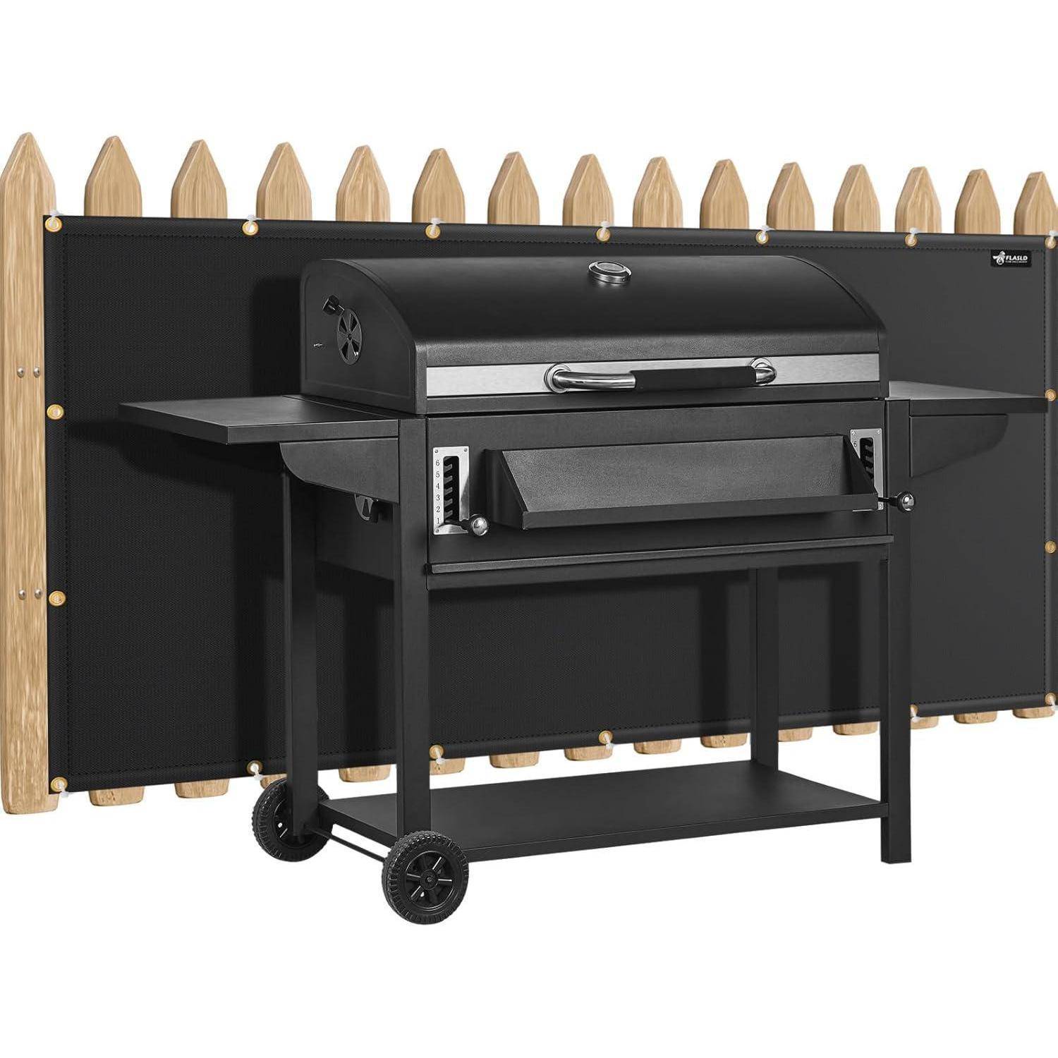 Ein schwarzer Barbecue-Grill mit Seitenablagen und Rädern steht vor einem Holzzaun. Er ist bereit für das Kochen im Freien.