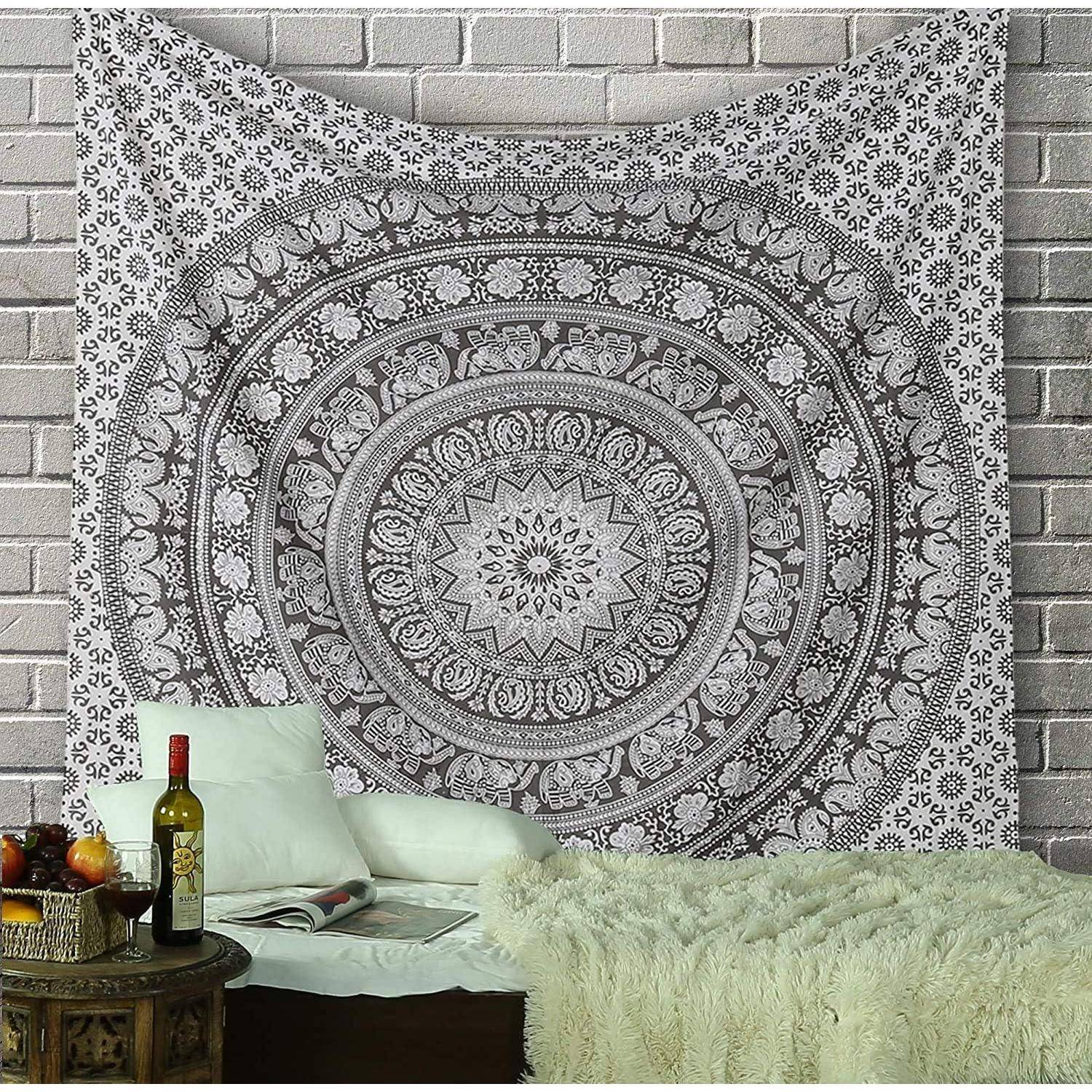 Ein Wandteppich mit kunstvollen Mandala-Mustern hängt an einer Ziegelwand über einem gemütlichen Sitzbereich mit Kissen, einer Decke, einer Weinflasche und Obst.