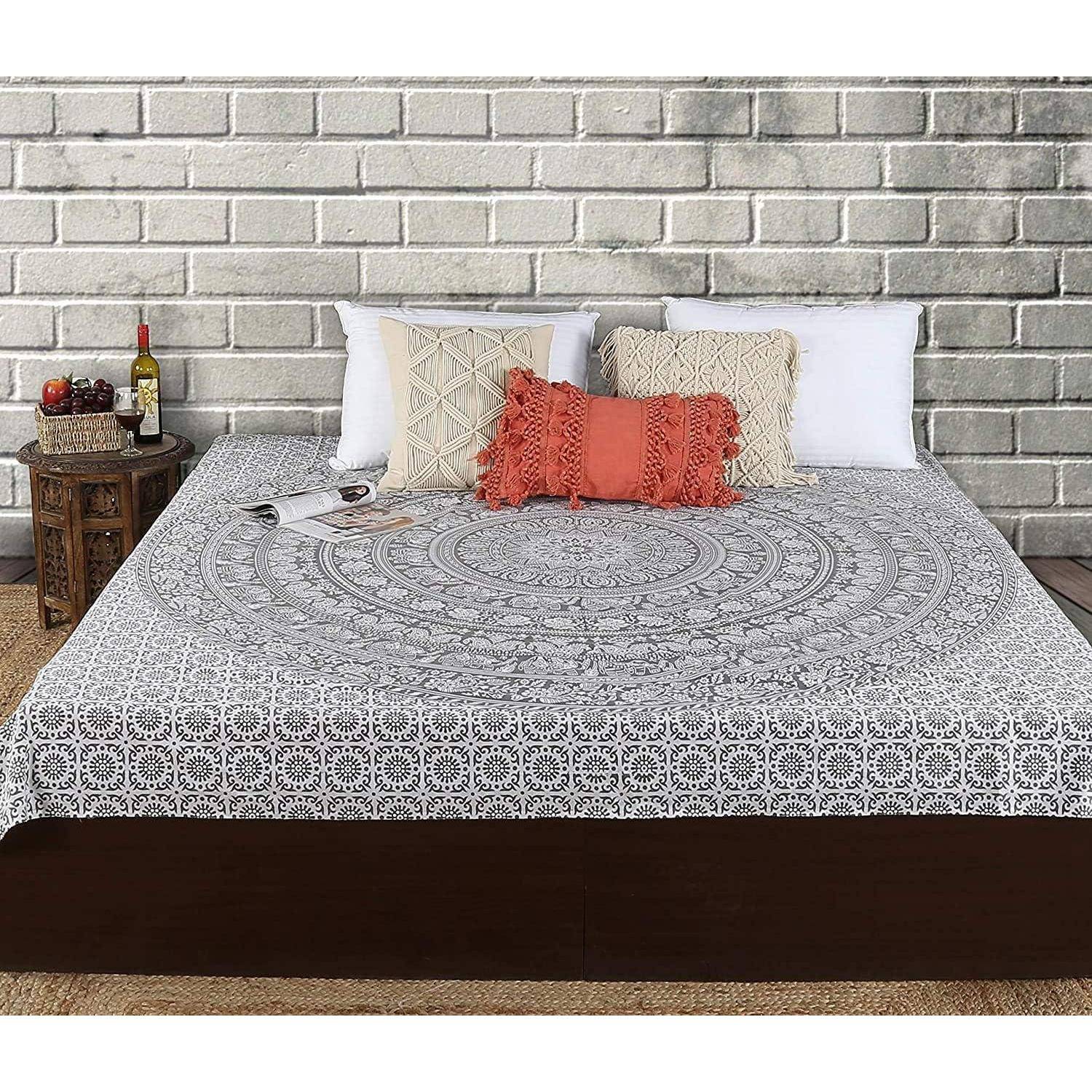 Ein Bett mit einem grau-weiß gemusterten Mandala-Bettüberwurf, zwei weißen Kissen und einem orangefarbenen Zierkissen. Ein Nachttisch trägt Deko und eine Weinflasche.