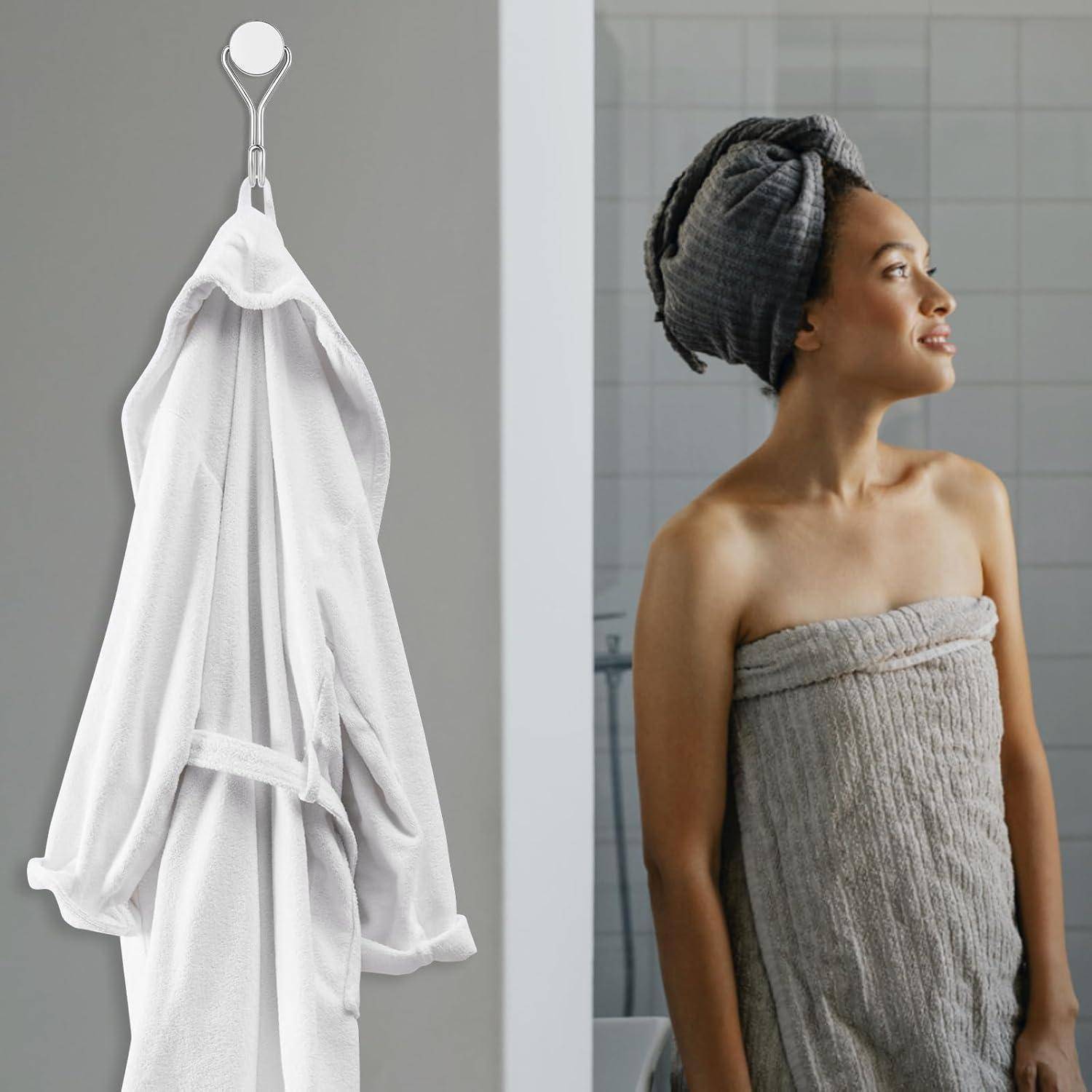 Eine Person, in ein graues Handtuch und einen Turban gehüllt, lächelt und blickt zur Seite. Ein weißer Bademantel hängt an einem Haken neben ihr.