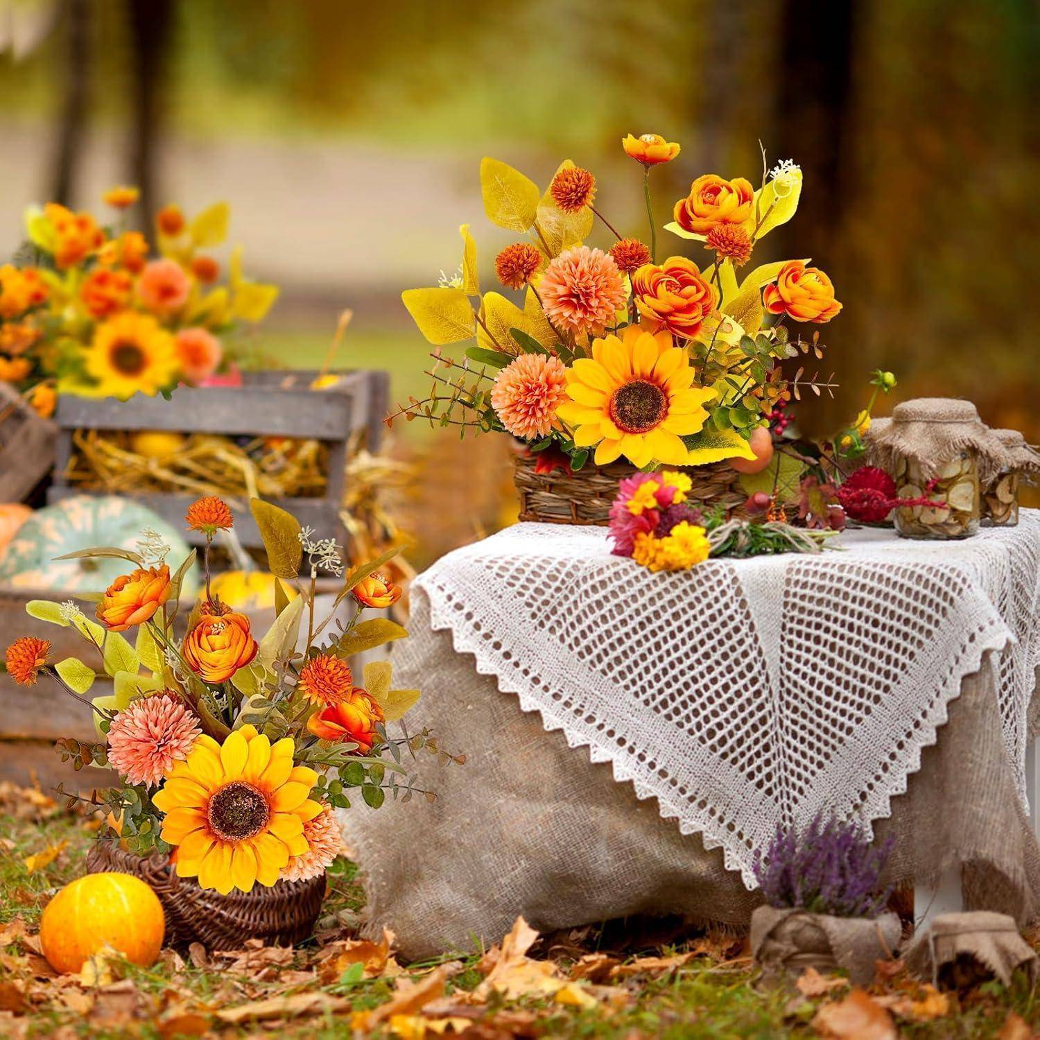 Eine rustikale Herbst-Außenauslage mit Körben voller Sonnenblumen und Chrysanthemen auf einem mit einem Tuch bedeckten Tisch, umgeben von herabgefallenen Blättern.