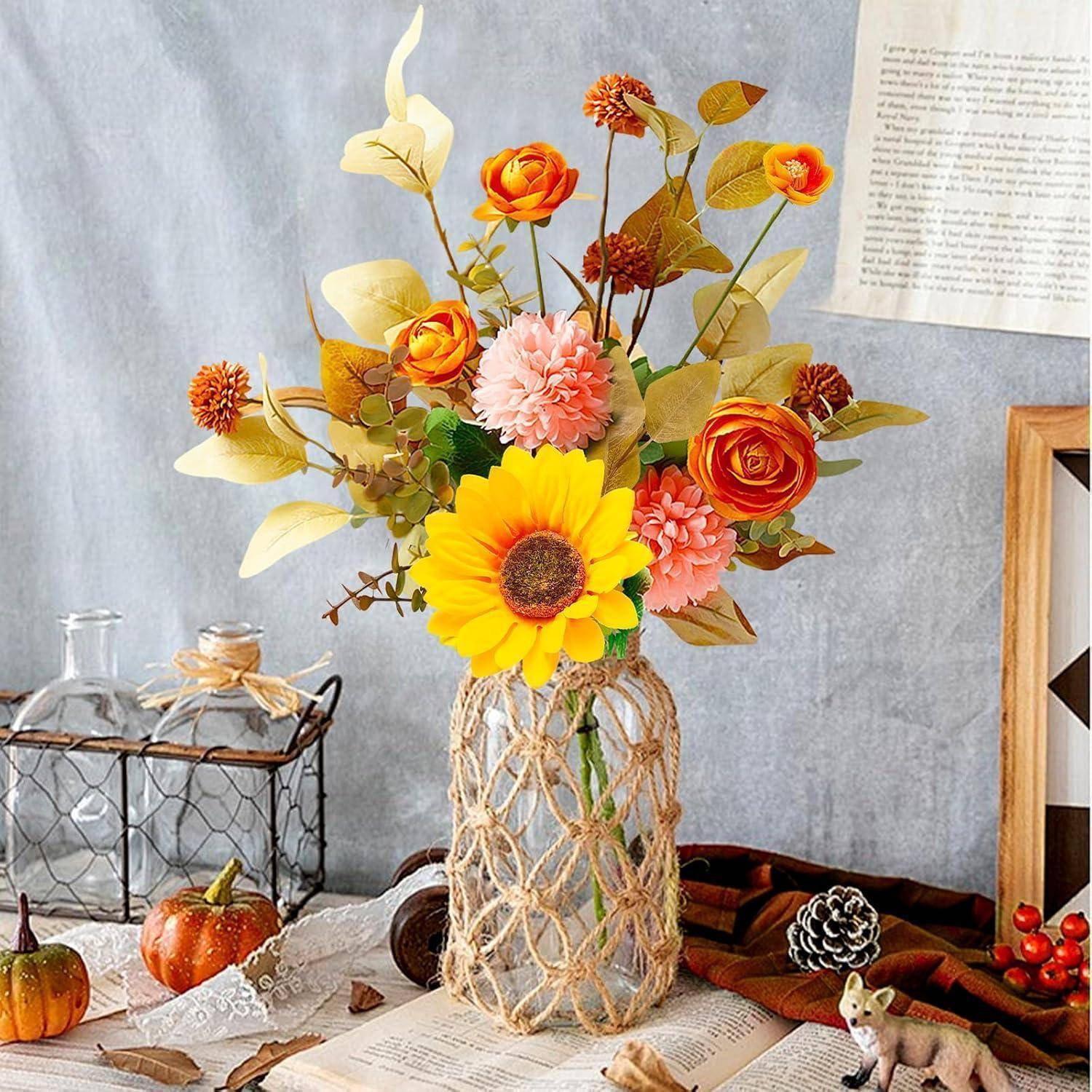 Eine Vase mit Herbstblumen, darunter eine leuchtend gelbe Sonnenblume, ist auf einem rustikalen Tisch neben Herbstblättern und einem kleinen Kürbis arrangiert.