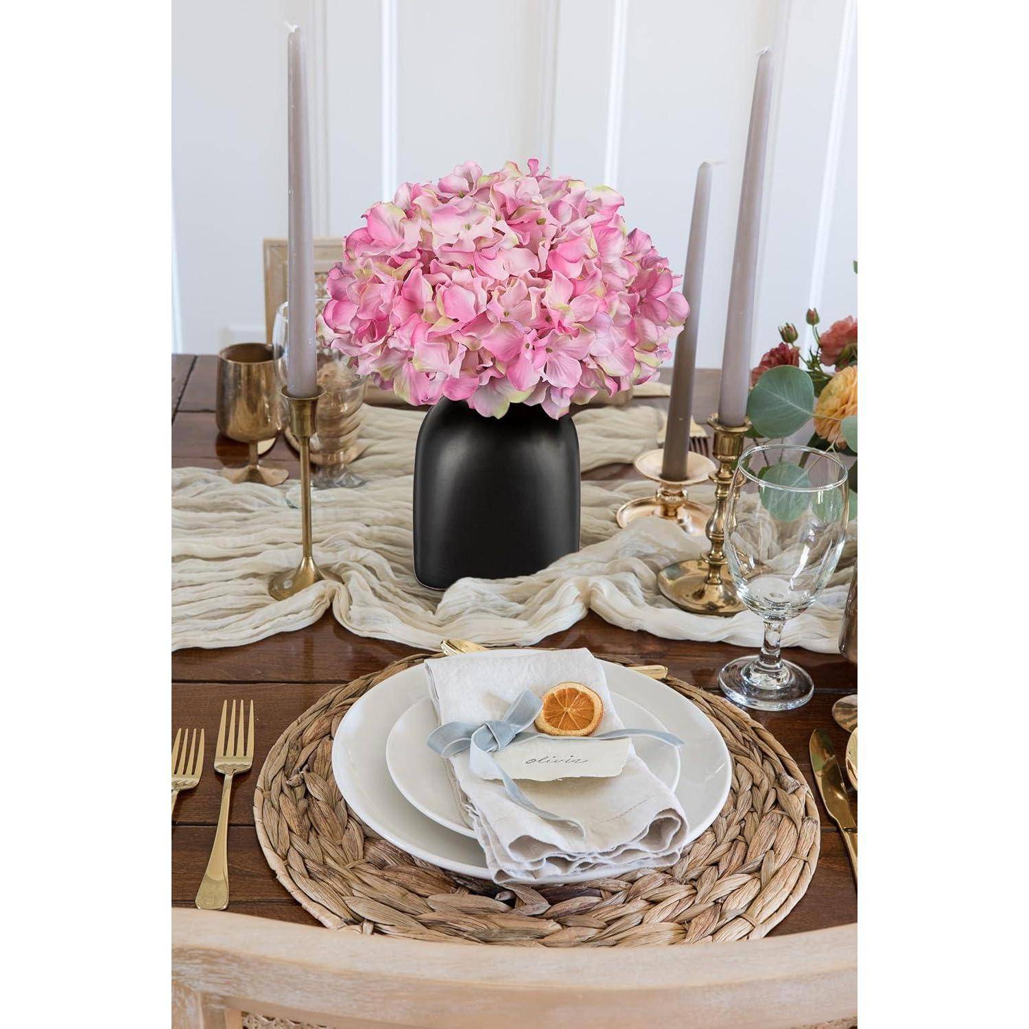 Ein Esstisch-Arrangement mit einem geflochtenen Tischset, weißem Teller, Stoffserviette mit getrockneter Orangenscheibe und rosa Blumen in einer schwarzen Vase als Mittelpunkt.