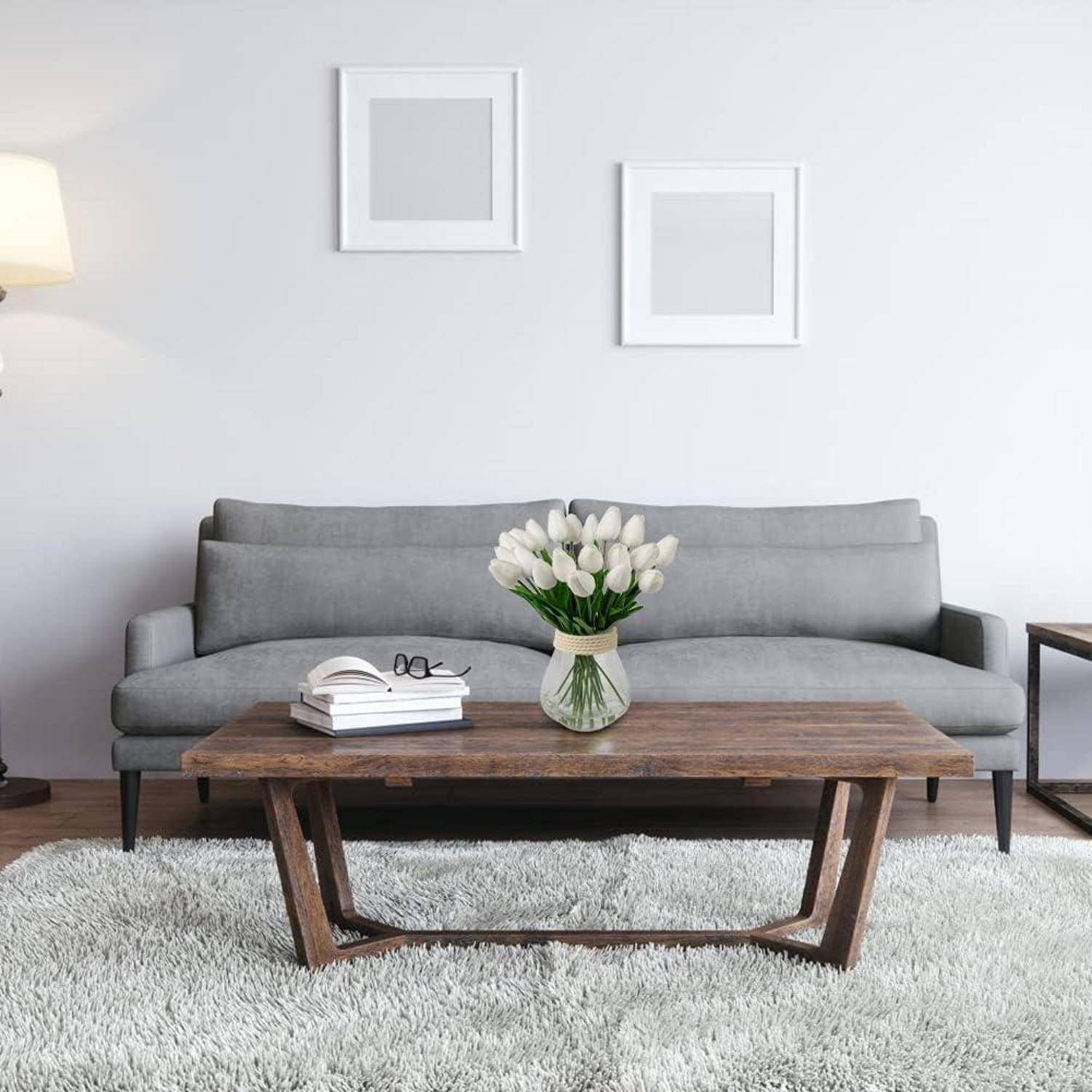 Ein modernes Wohnzimmer mit einem grauen Sofa, zwei quadratischen Rahmen an der Wand, einem Holzcouchtisch mit einem Buch und einer Vase mit weißen Tulpen auf einem hellen Teppich.