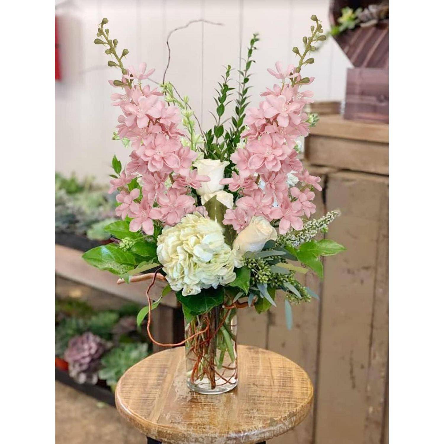 Eine Blumengestaltung mit rosa und weißen Blumen, grünen Blättern und verdrehten Zweigen in einer Glasvase auf einem Holzhocker.
