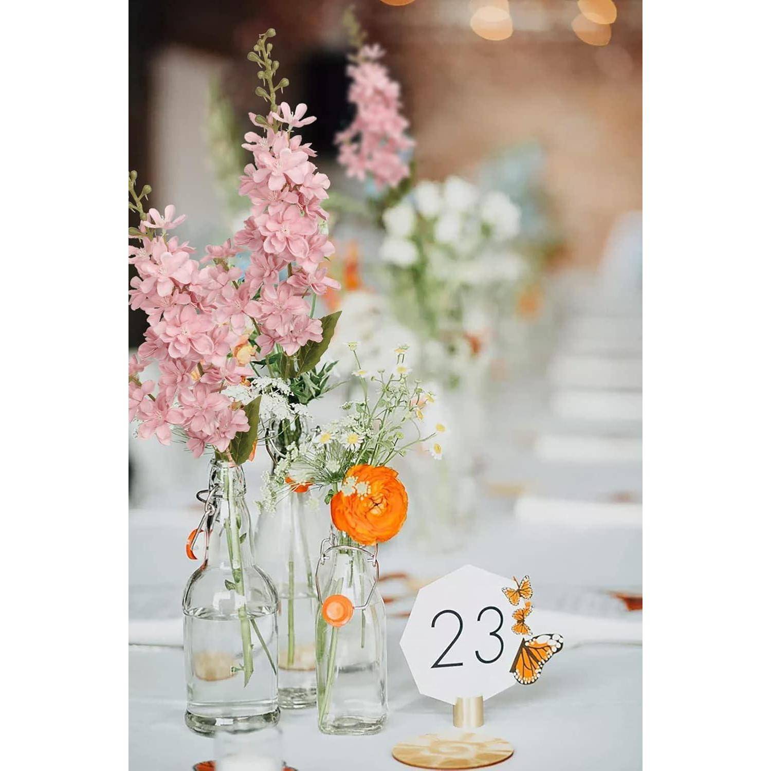 Tischdekoration mit rosa und weißen Blumen in Glasflaschen, hellen orangefarbenen Akzenten und einer Tischnummerkarte mit der Zahl '23' und Schmetterlingen.
