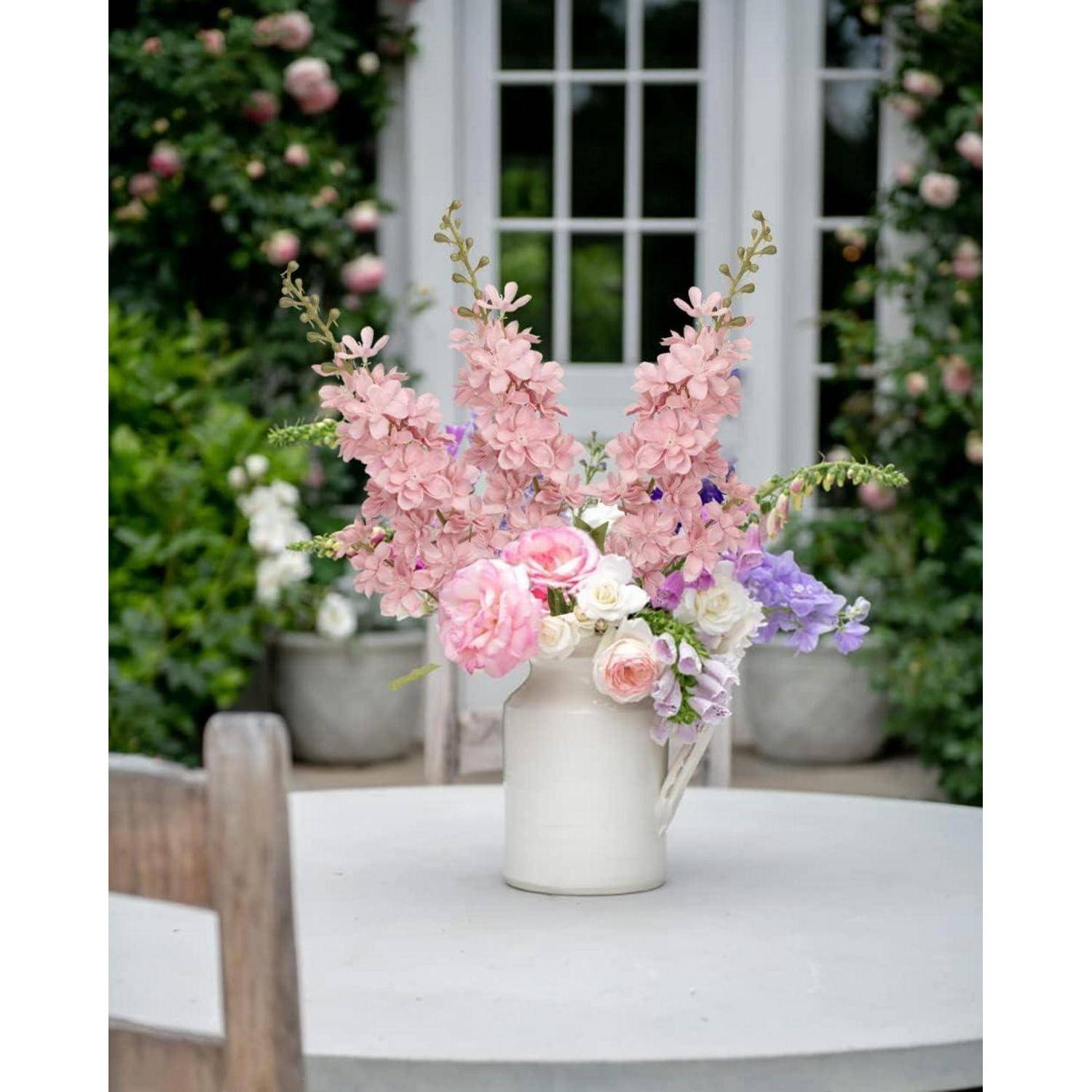 Eine Vase mit rosa und violetten Blumen auf einem Tisch, vor einer Gartenkulisse mit einer teilweise sichtbaren Tür und grünem Blattwerk.