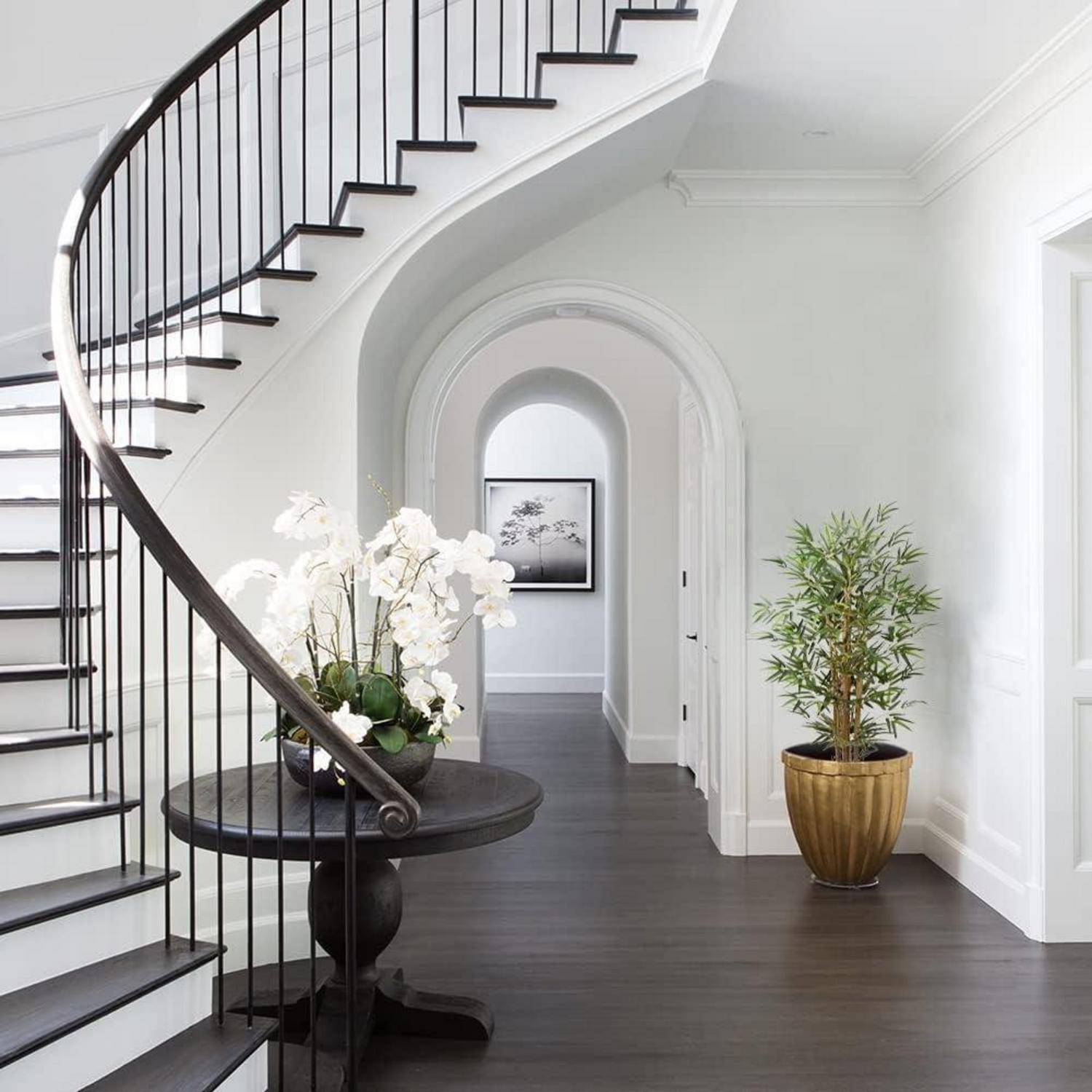 Elegante Diele mit einer geschwungenen Treppe, dunklem Bodenleger, weißen Wänden, einem runden Tisch mit weißen Orchideen und einer Topfpflanze in der Nähe eines Torbogens.