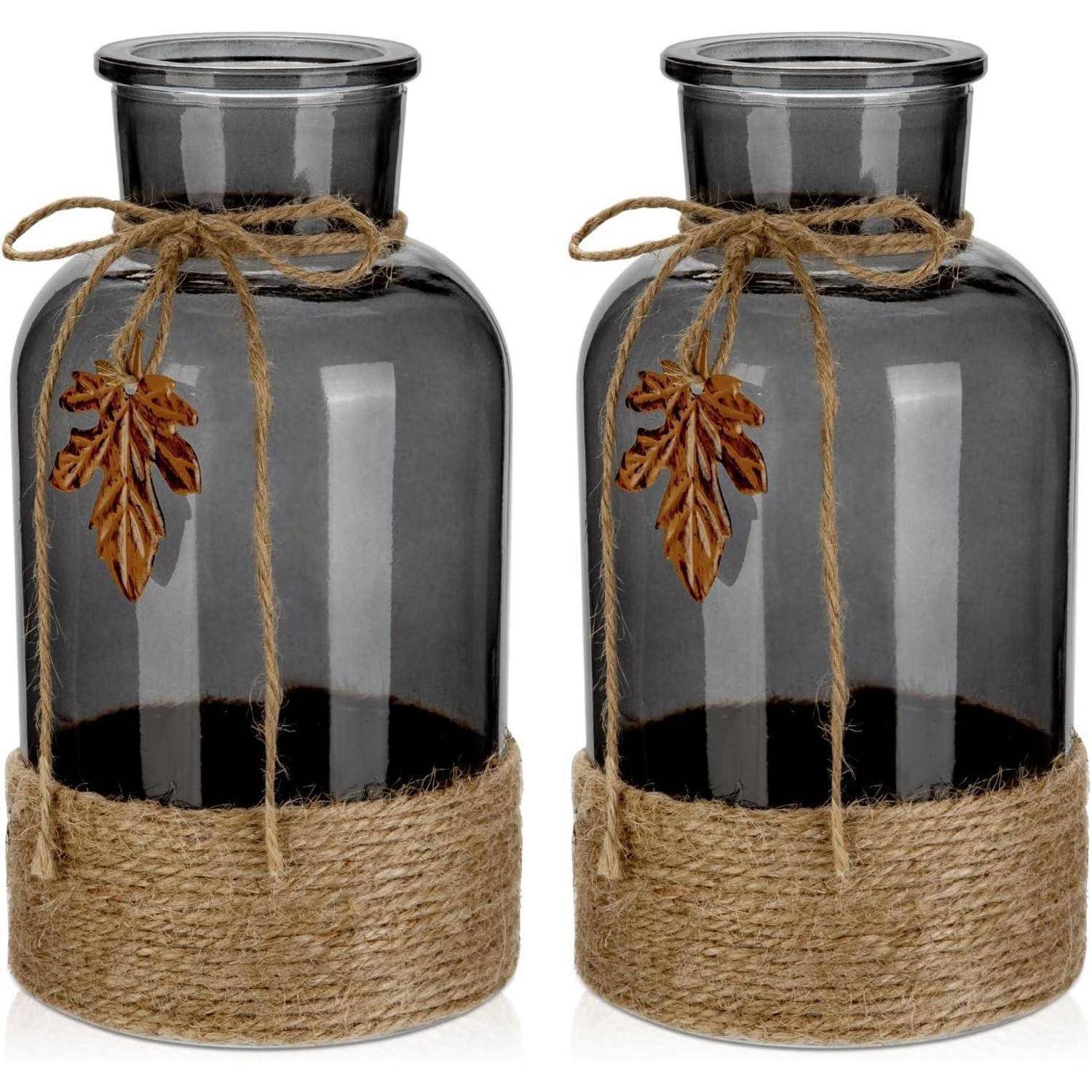 Zwei Glasgefäße mit Juteseilen als Akzente und Blattornamenten. Die Gläser sind am Hals zusammengebunden und haben ein rustikales Design.