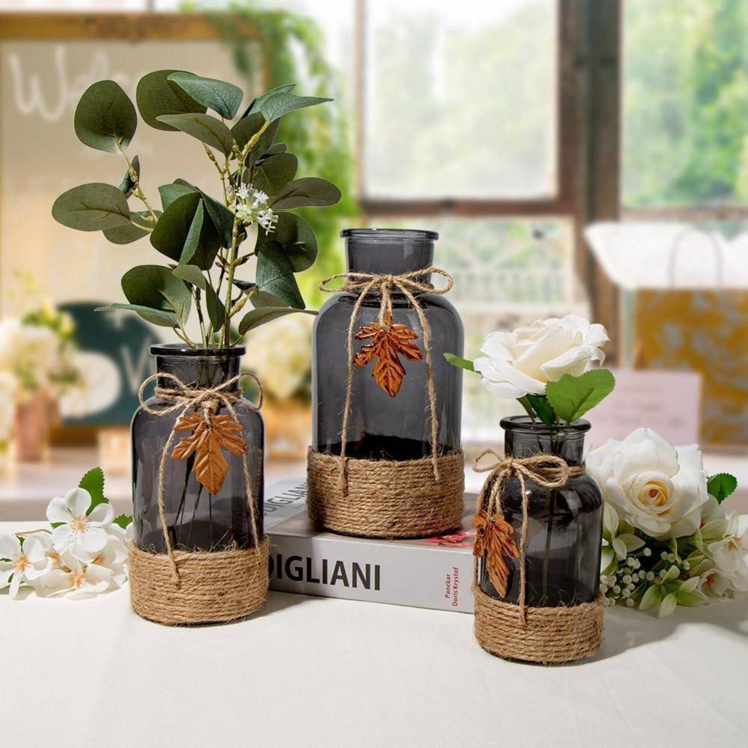 Drei dunkle Glasvasen, umwickelt mit Schnur, geschmückt mit Blättern und gefüllt mit weißen Blumen, stehen auf einem Tisch in der Nähe eines Fensters.