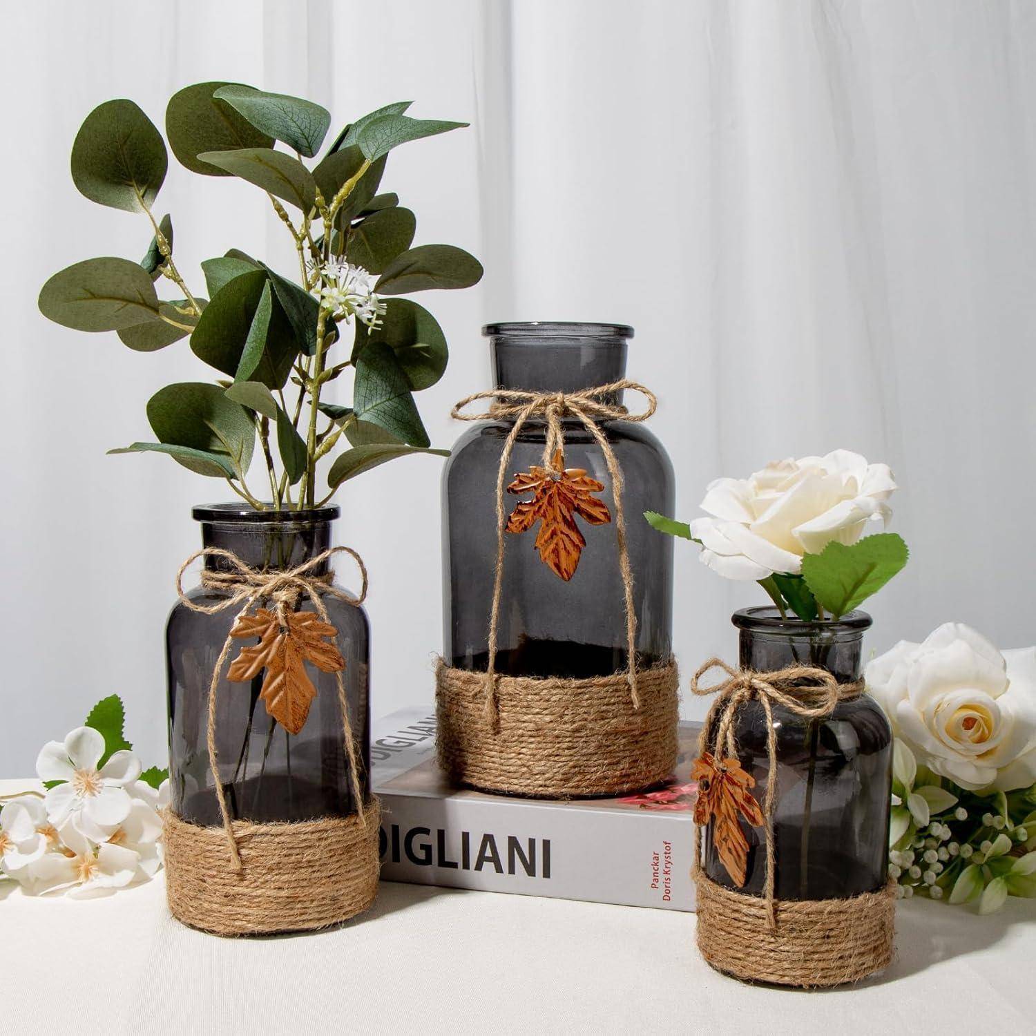 Drei dunkle Glasvasen, umwickelt mit Bindfaden und geschmückt mit Blättern und weißen Blumen, angeordnet auf einem Buch. Zusätzliche Blumen umgeben die Anordnung.