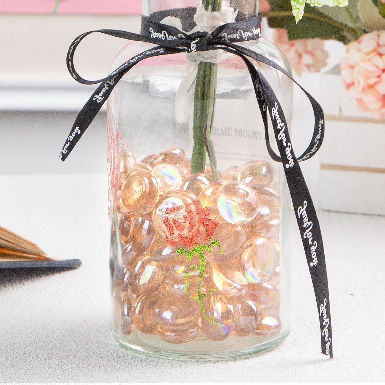 Eine Glasvase mit dekorativen Steinen und einer einzelnen Rose im Inneren, mit einem schwarzen Band mit weißer Schrift umwickelt, steht auf einem Tisch.