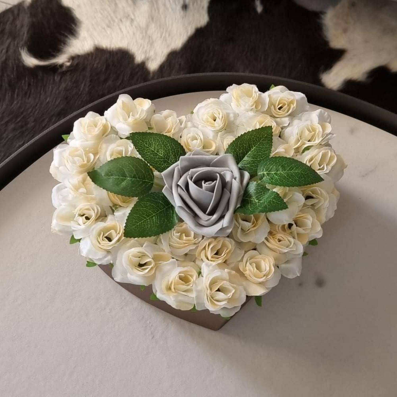 Herzförmige Blumenanordnung mit weißen Rosen, die eine zentrale graue Rose umgeben, vor einem schwarz-weißen Fellhintergrund.