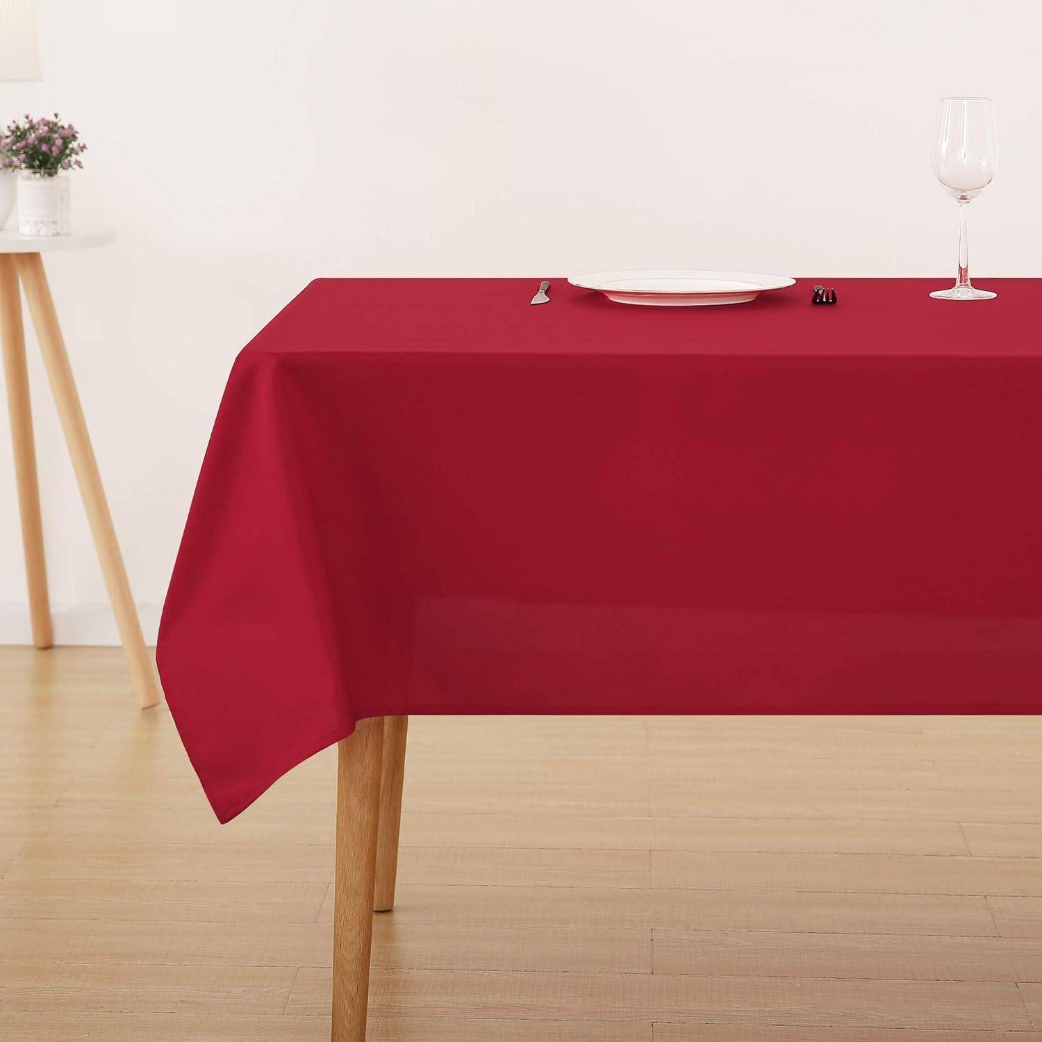 Ein rechteckiger Esstisch mit einer roten Tischdecke ist ordentlich gedeckt mit einem weißen Teller, Besteck und einem leeren Weinglas vor einem hellen Hintergrund.